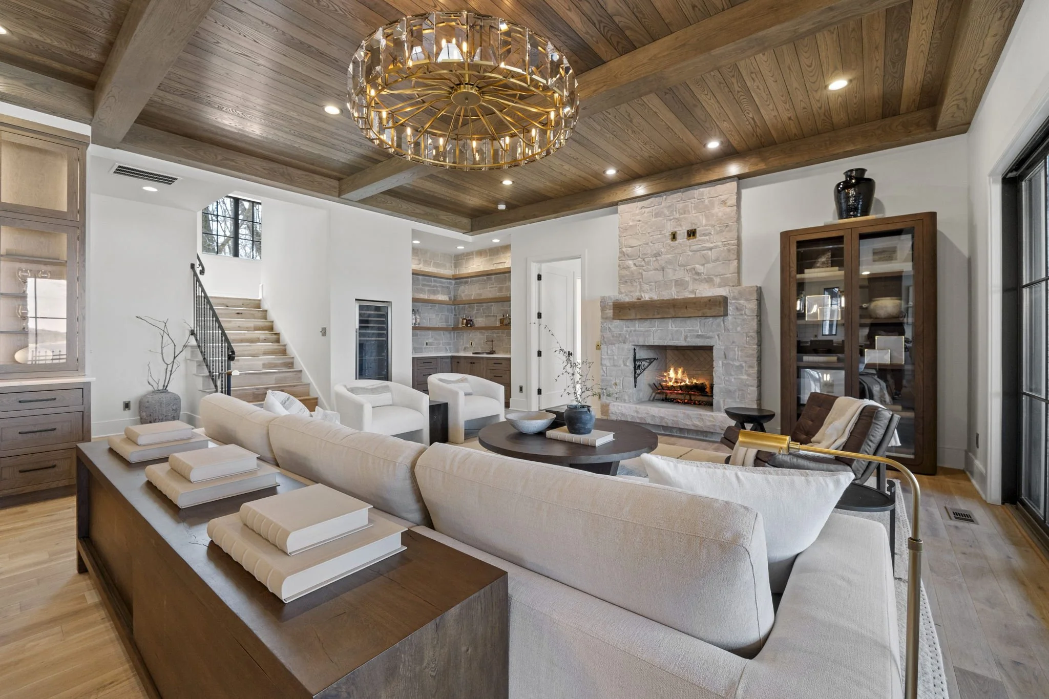 Living room with a stone fireplace, wooden ceiling with a chandelier, white sofa, two white armchairs, a round wooden coffee table, and a wooden cabinet. There's a sliding glass door on the right and stairs leading up on the left.