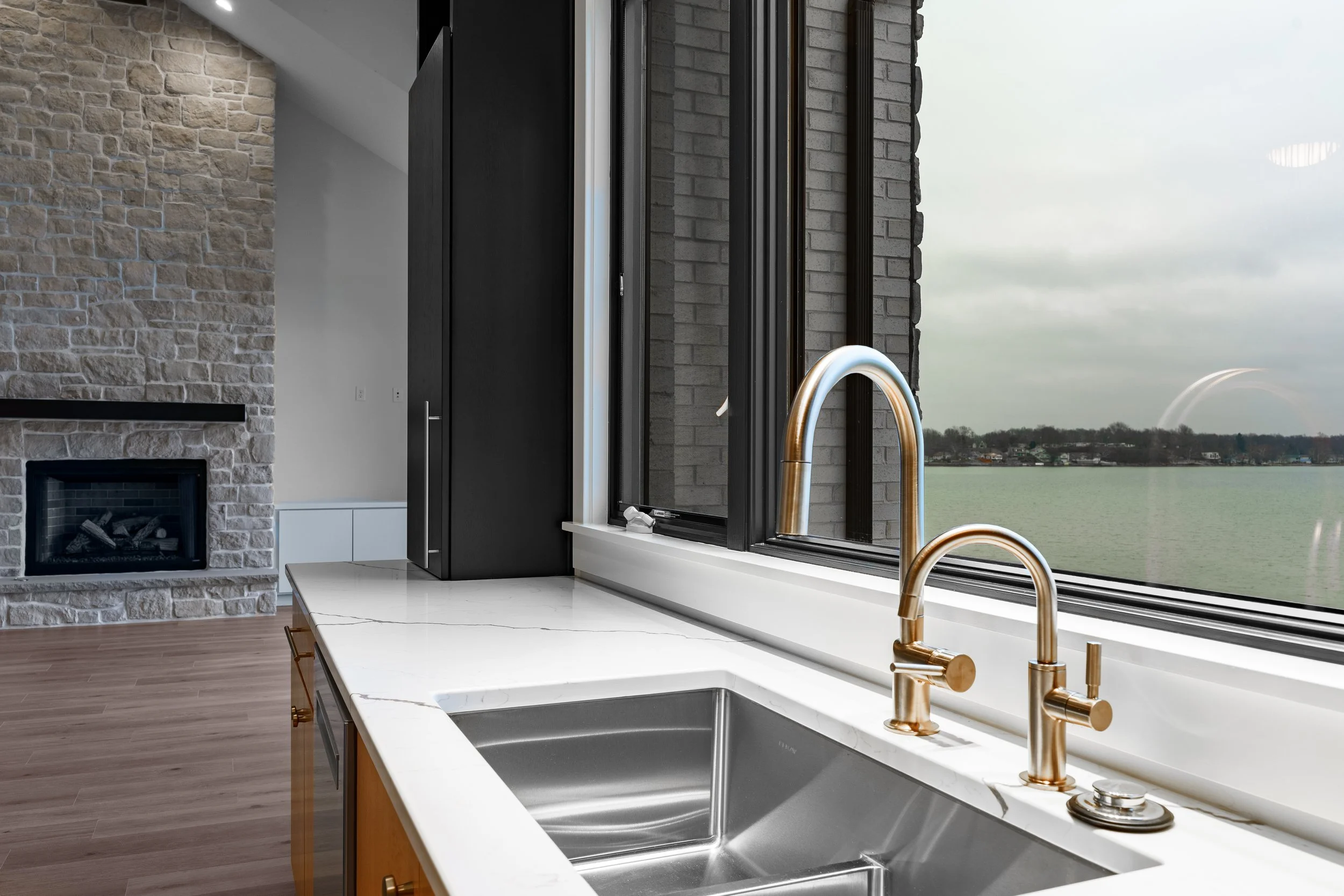 Modern kitchen with white marble countertops, gold sink and faucet, large window overlooking water, and a stone fireplace in the living room area.