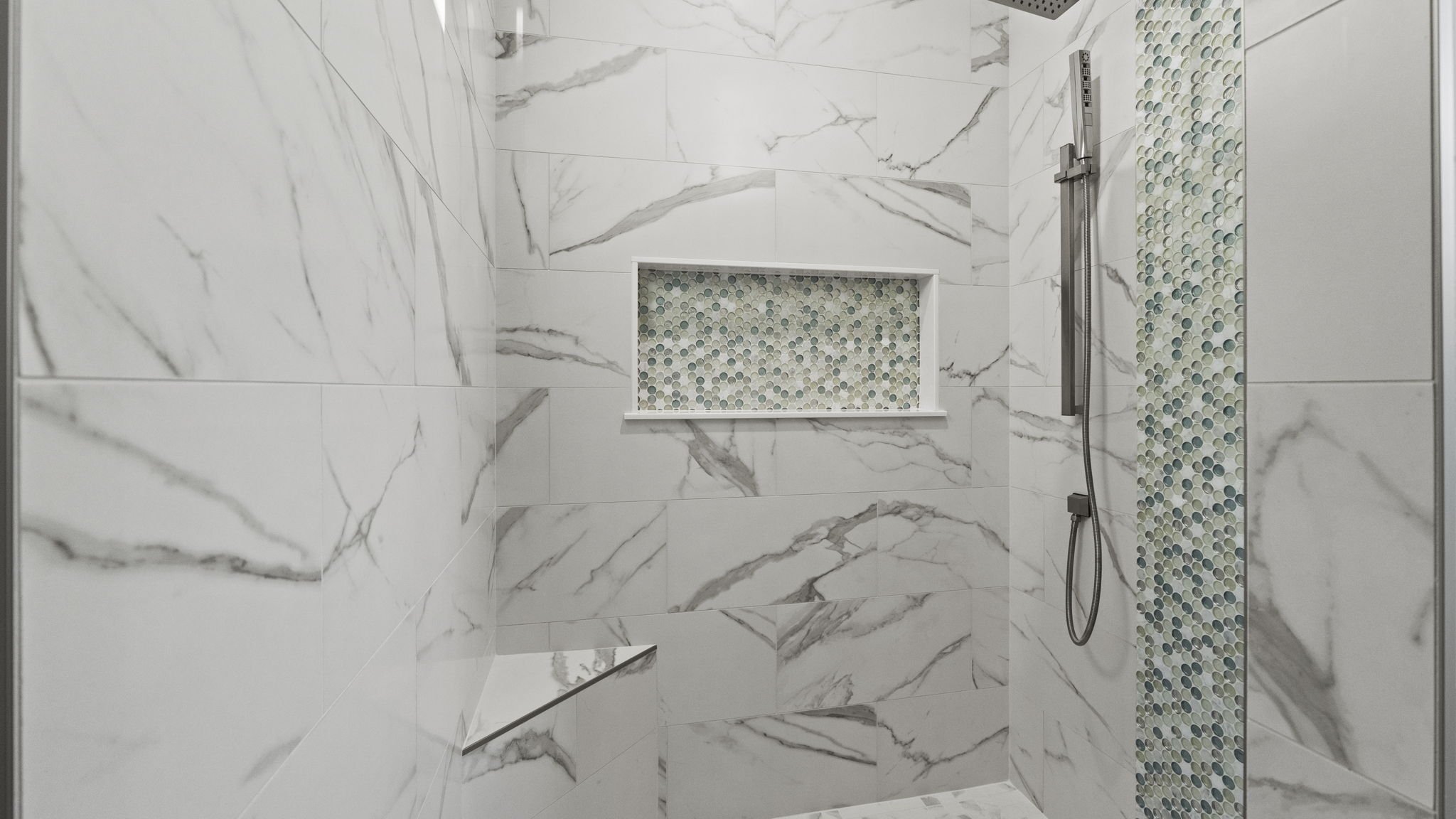 A modern walk-in shower with white marble tiles and gray veining, a built-in shelf with mosaic tile backing, and a handheld showerhead mounted on the wall.