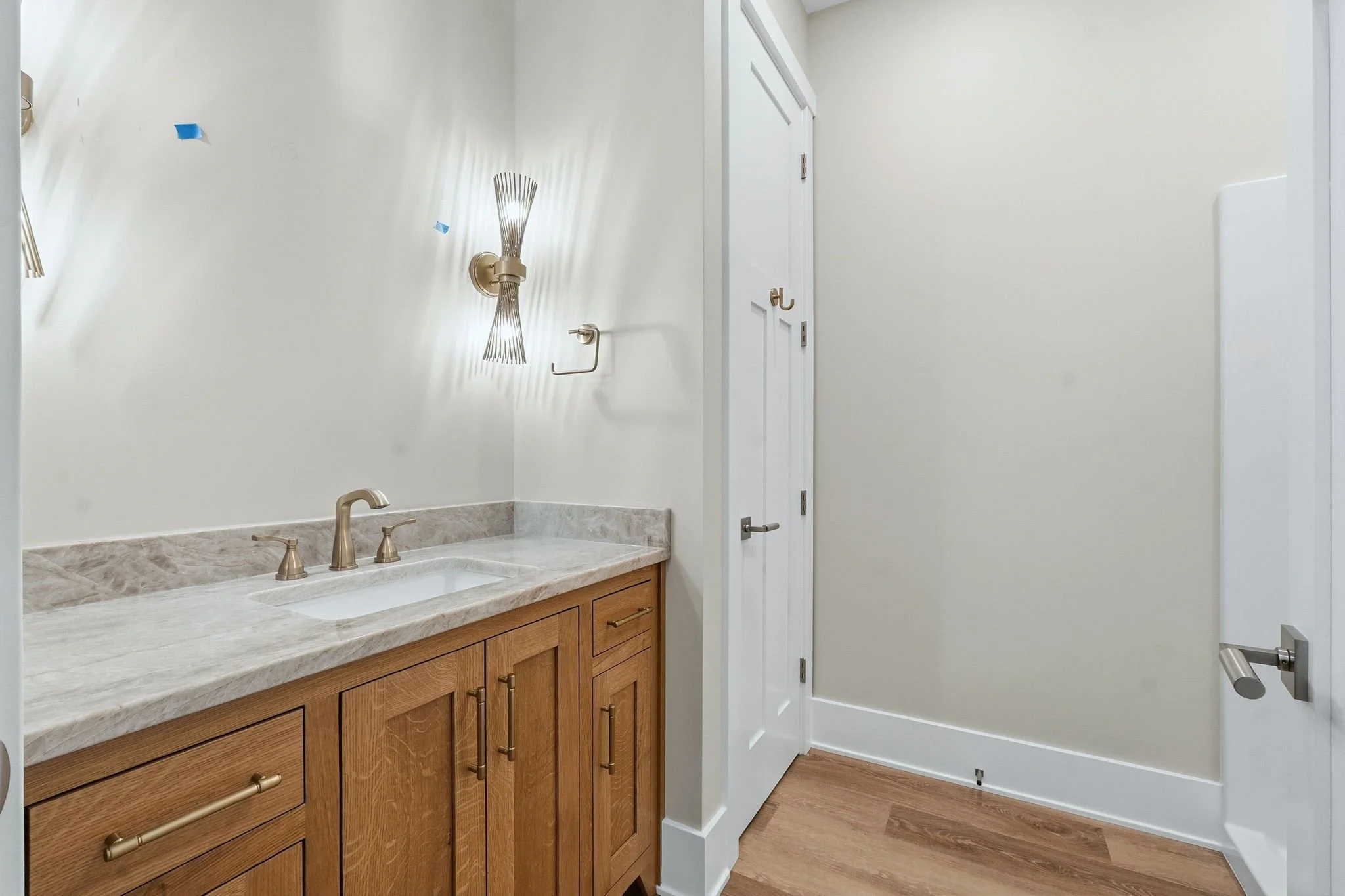 Bathroom with wooden cabinet and marble countertop, gold fixtures, white walls, wood flooring, and wall-mounted lamps