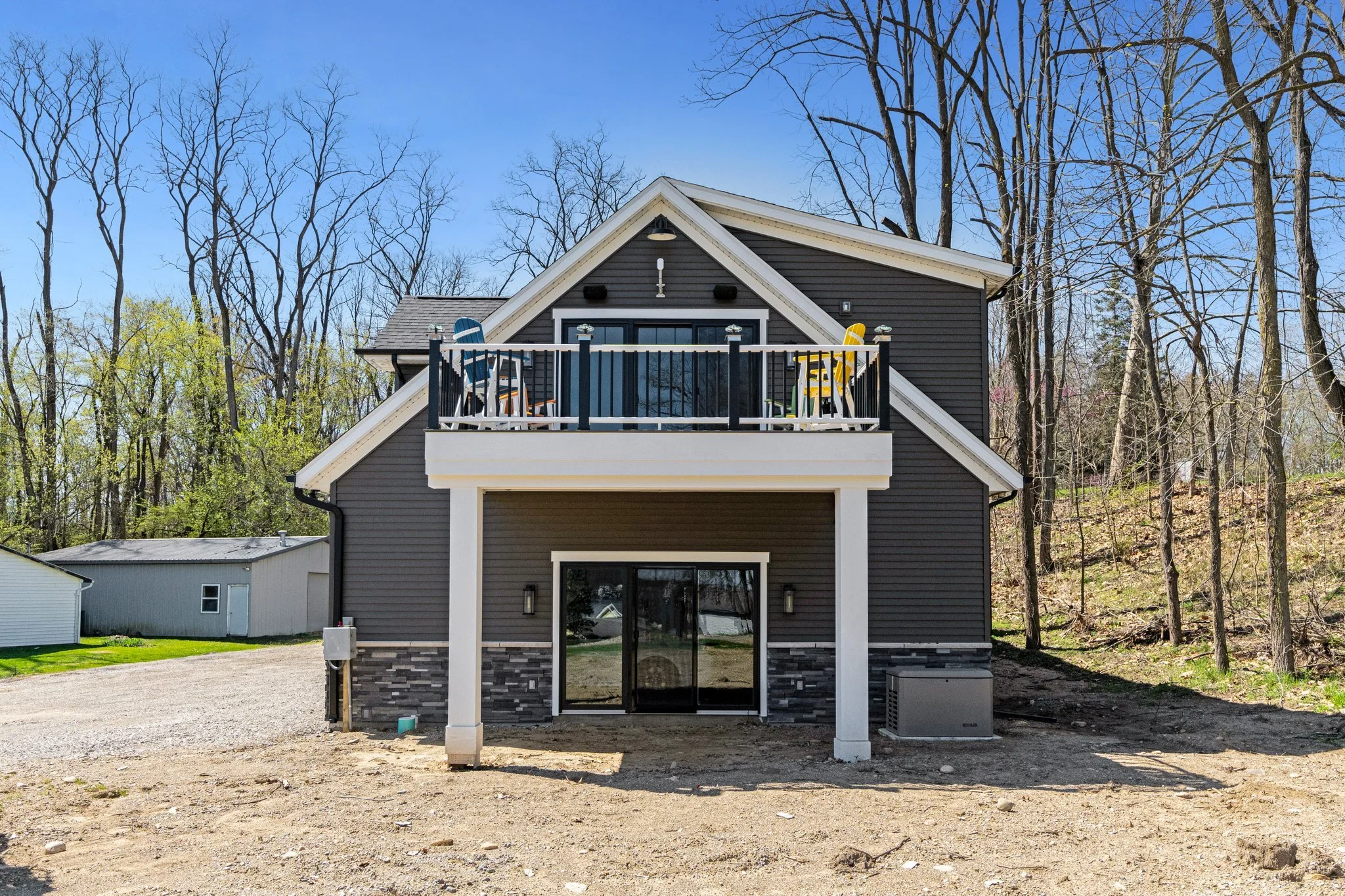 Modern two-story house with dark siding, a balcony with colorful chairs, surrounded by leafless trees, with gravel and dirt yard in foreground.