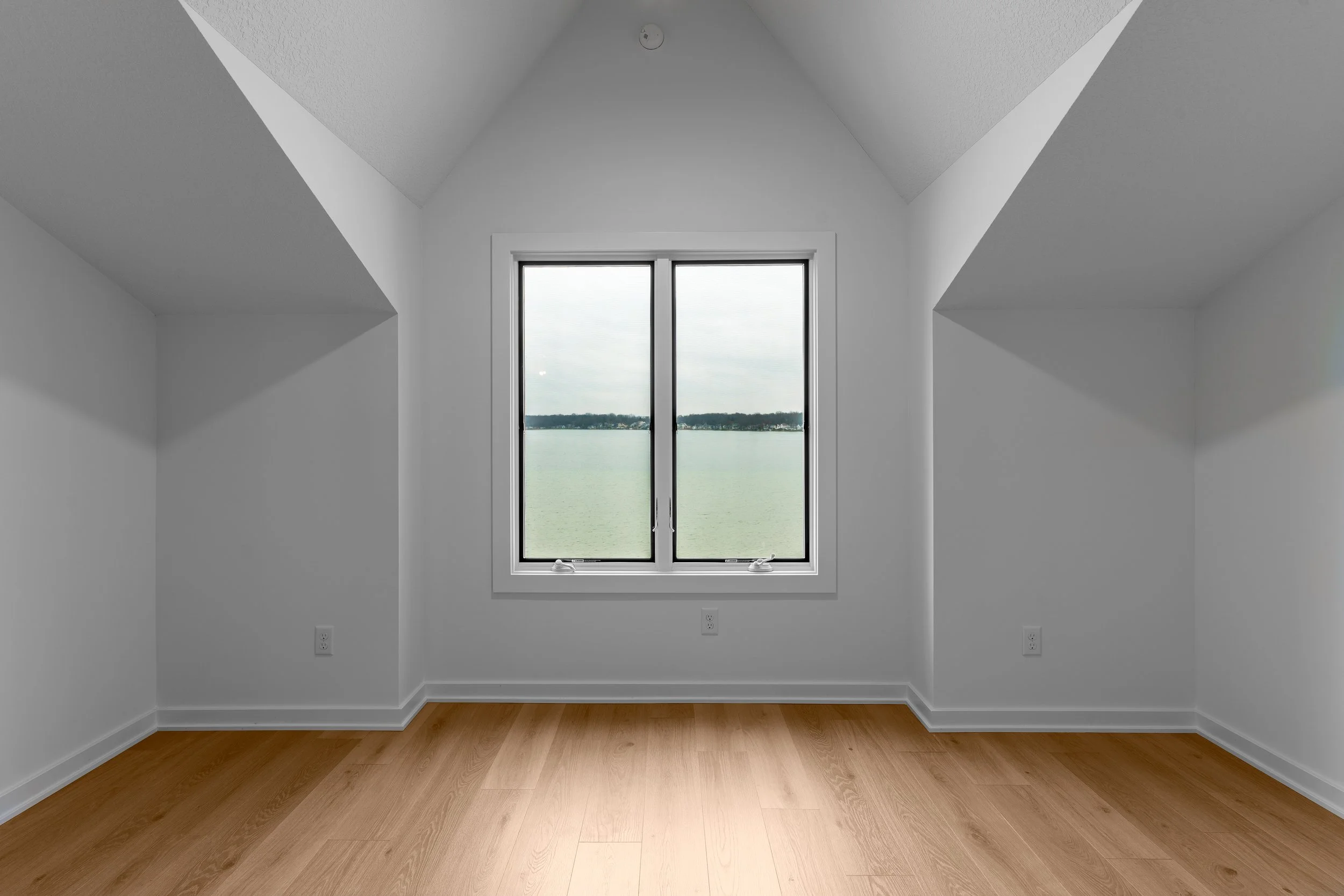 Empty white room with sloped ceilings and a large window overlooking water and sky, with wooden floor and electrical outlets.