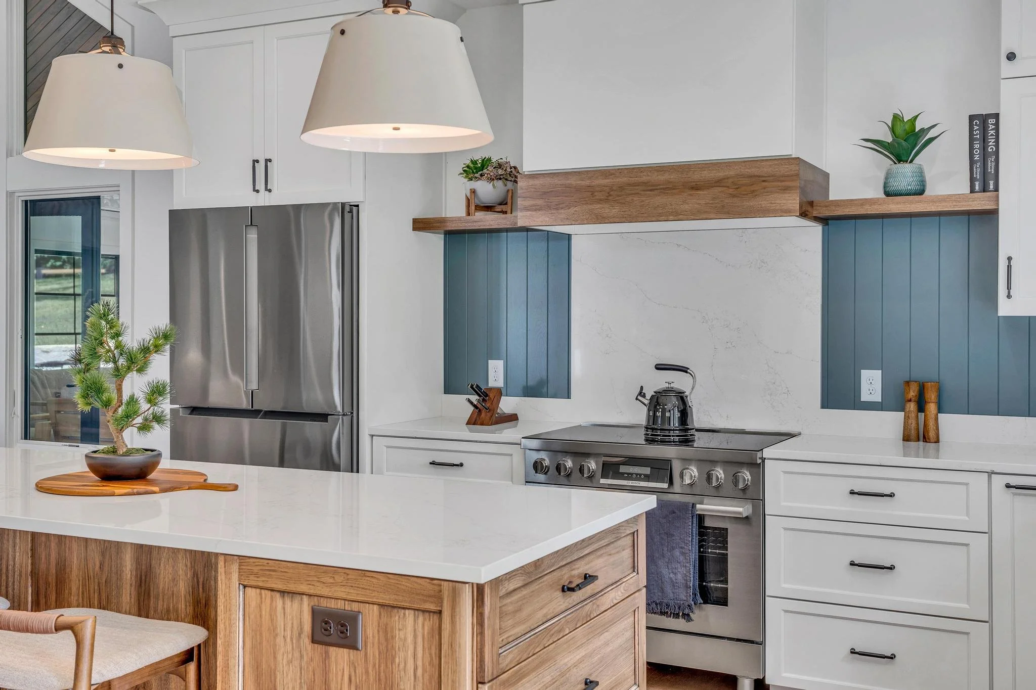 Modern kitchen featuring a white island with a potted plant, stainless steel refrigerator, stove with a black kettle, wooden and blue accents, and decorative plants on open shelves.