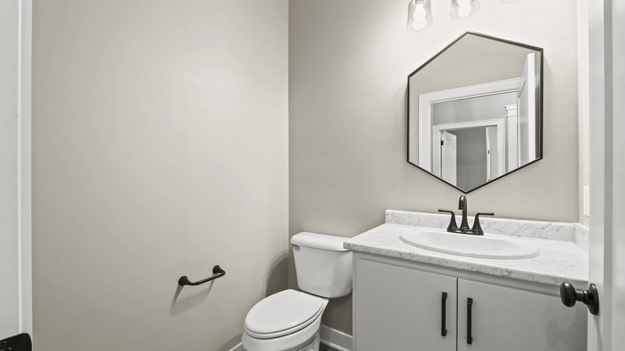 A small bathroom with a toilet, a white vanity with a marble countertop, a black faucet, and a large wall mirror. The walls are painted light beige and there is a towel bar on the wall next to the toilet.