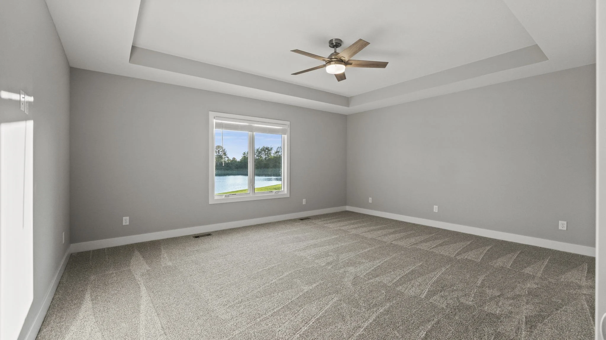 Empty bedroom with beige carpet, gray walls, a white-trimmed window showing a lake and green trees outside, and a ceiling fan with a light and wooden blades.