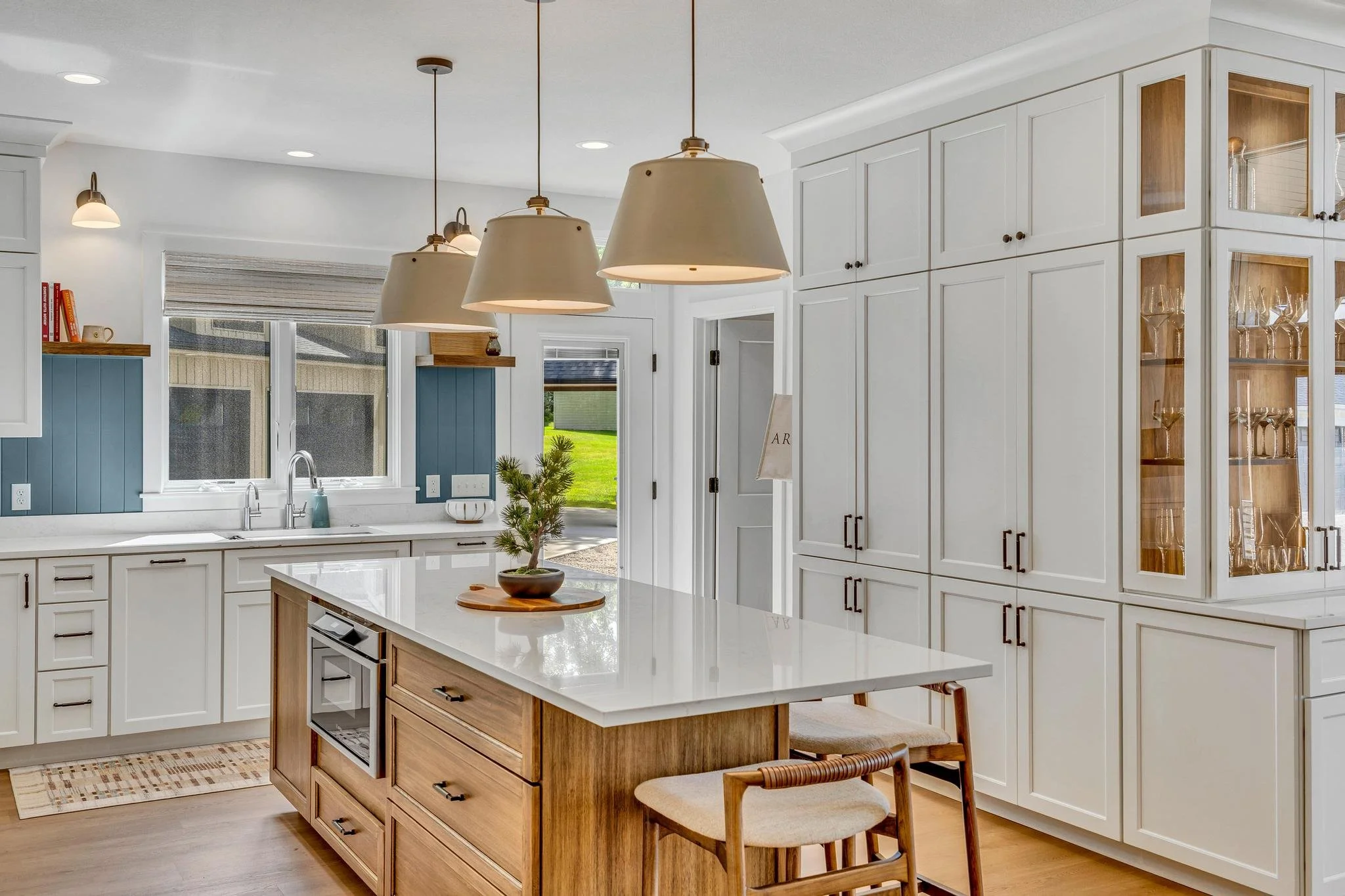 Modern kitchen with white cabinets, a large island with a wooden base, and pendant lighting. There are windows with a view of a green lawn outside, and a built-in glass cabinet showing glassware.