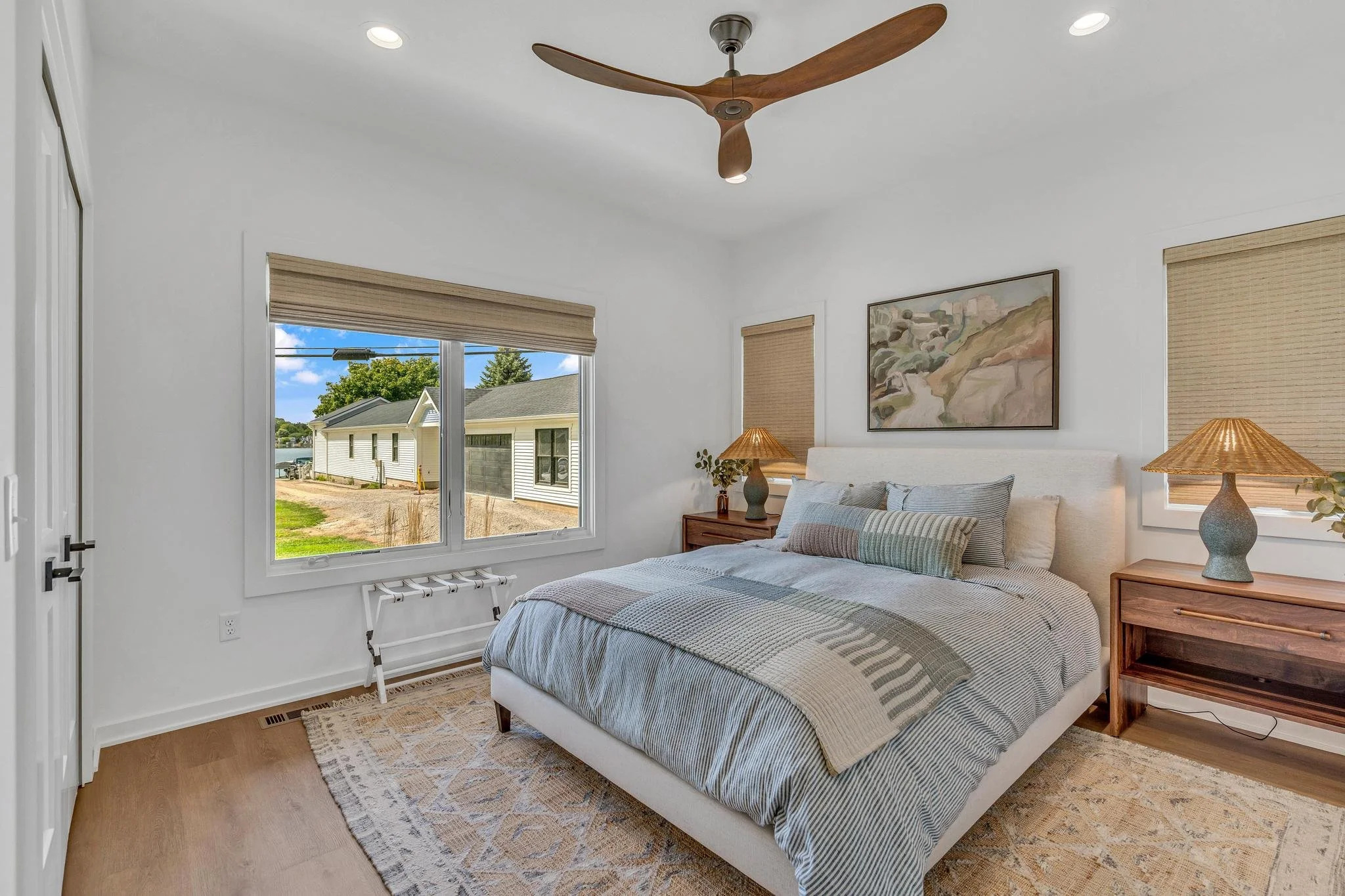 A bright bedroom with a large window showing a view of neighboring houses and trees, a white bed with gray and beige bedding, wooden side tables with lamps, and a ceiling fan, with framed artwork on the wall.