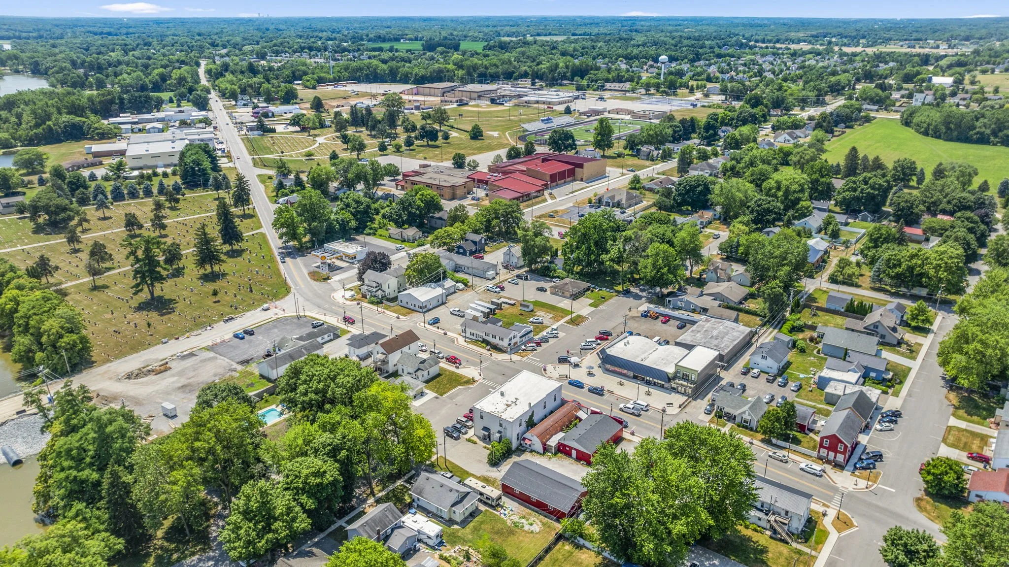 Aerial view of a small town with residential homes, commercial buildings, green trees, and open grassy areas under a partly cloudy sky.
