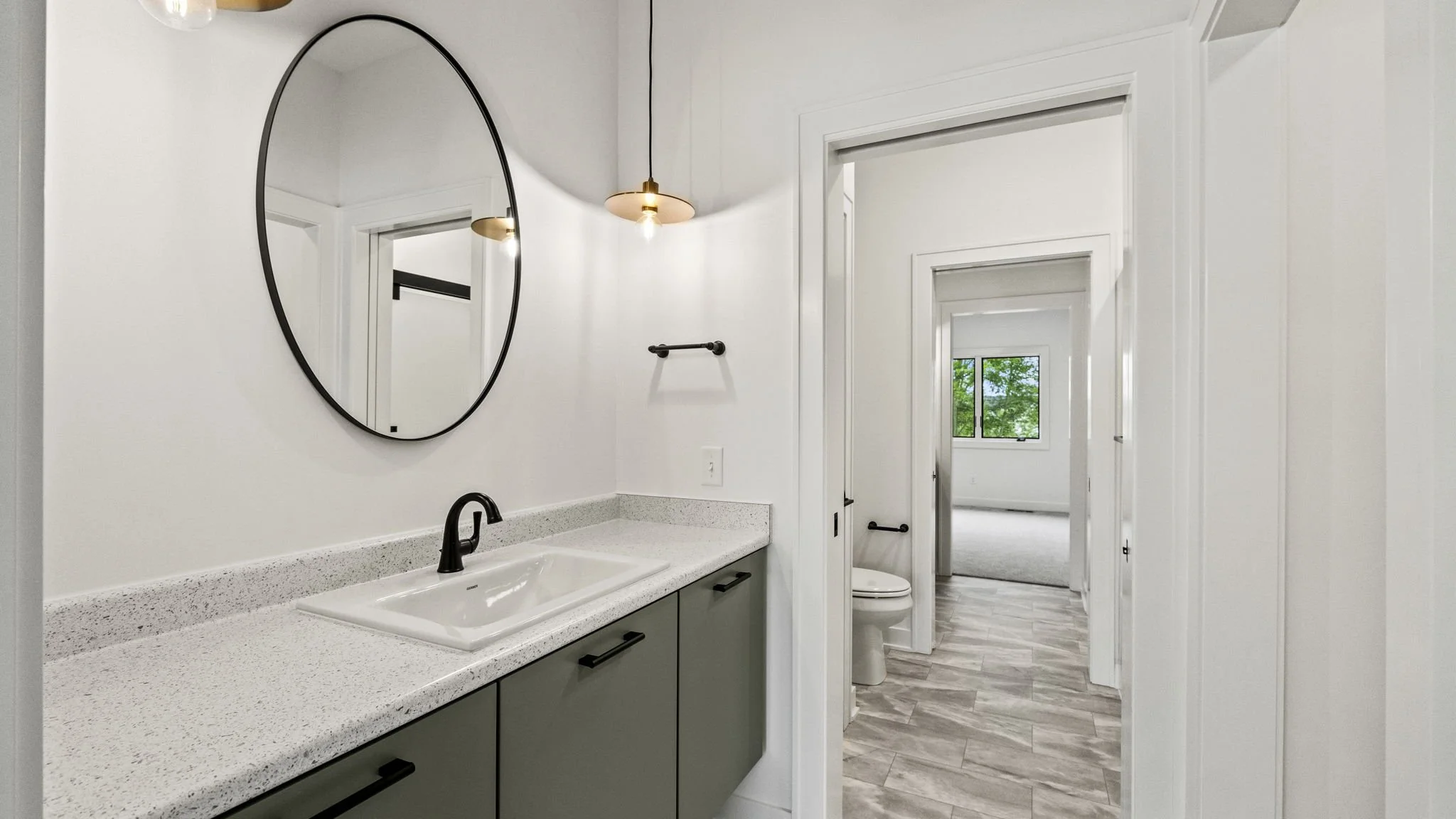 Bathroom with a black-framed oval mirror, white walls, a white sink with black faucet, green vanity cabinet, black towel bar, pendant light, and window visible through an open door leading to a bedroom.