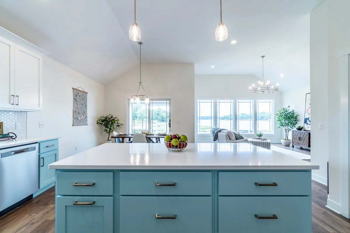 Bright, modern open-concept living space with a kitchen island, dining area, and living room, featuring large windows overlooking a body of water, with light-colored walls, hardwood floors, and contemporary lighting fixtures.