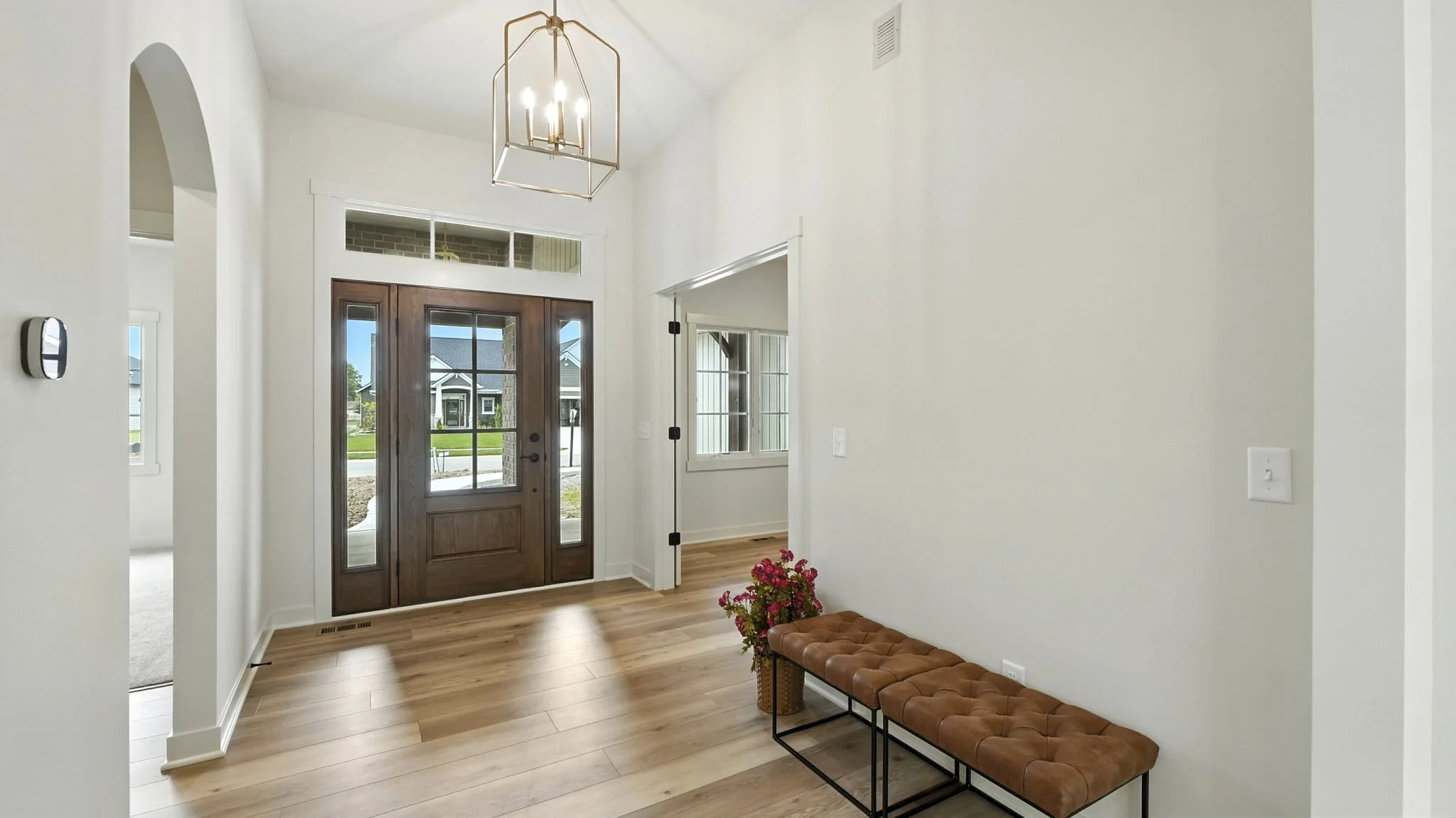 Entryway with wooden door and glass panels, wooden flooring, small bench with tufted brown seat, potted pink flowers, white walls, chandelier, window with blinds, and view of neighborhood outside.