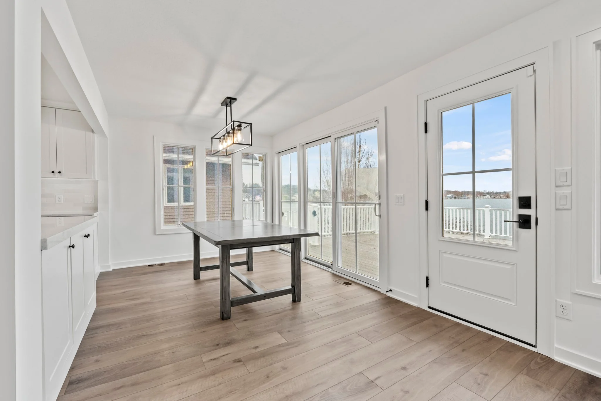 Bright dining area with large windows and glass door leading outside, empty wooden table in the center, white cabinets and sliding glass doors showing a view of water and outdoor deck.