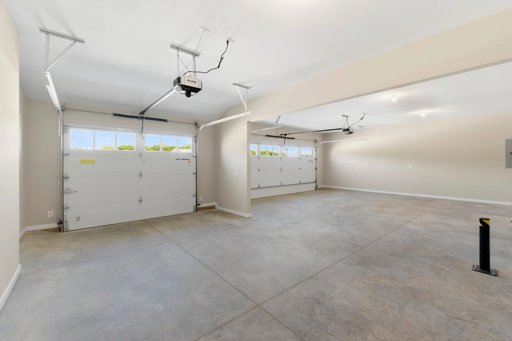 Empty two-car garage with concrete floor, white walls, and two white garage doors with windows; garage door openers attached to ceiling; natural light coming through windows; a black and yellow safety post on the right.