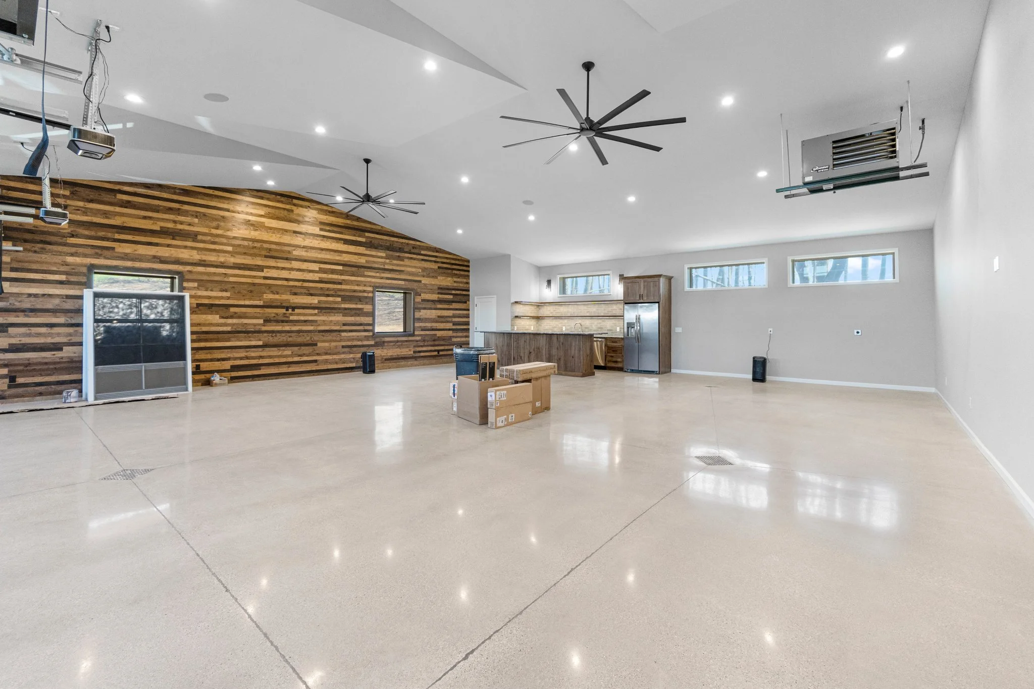 An empty, spacious room with polished concrete floors, white walls, multiple ceiling lights, black ceiling fans, a wooden accent wall, and a modern kitchen with stainless steel appliances and wooden cabinetry.