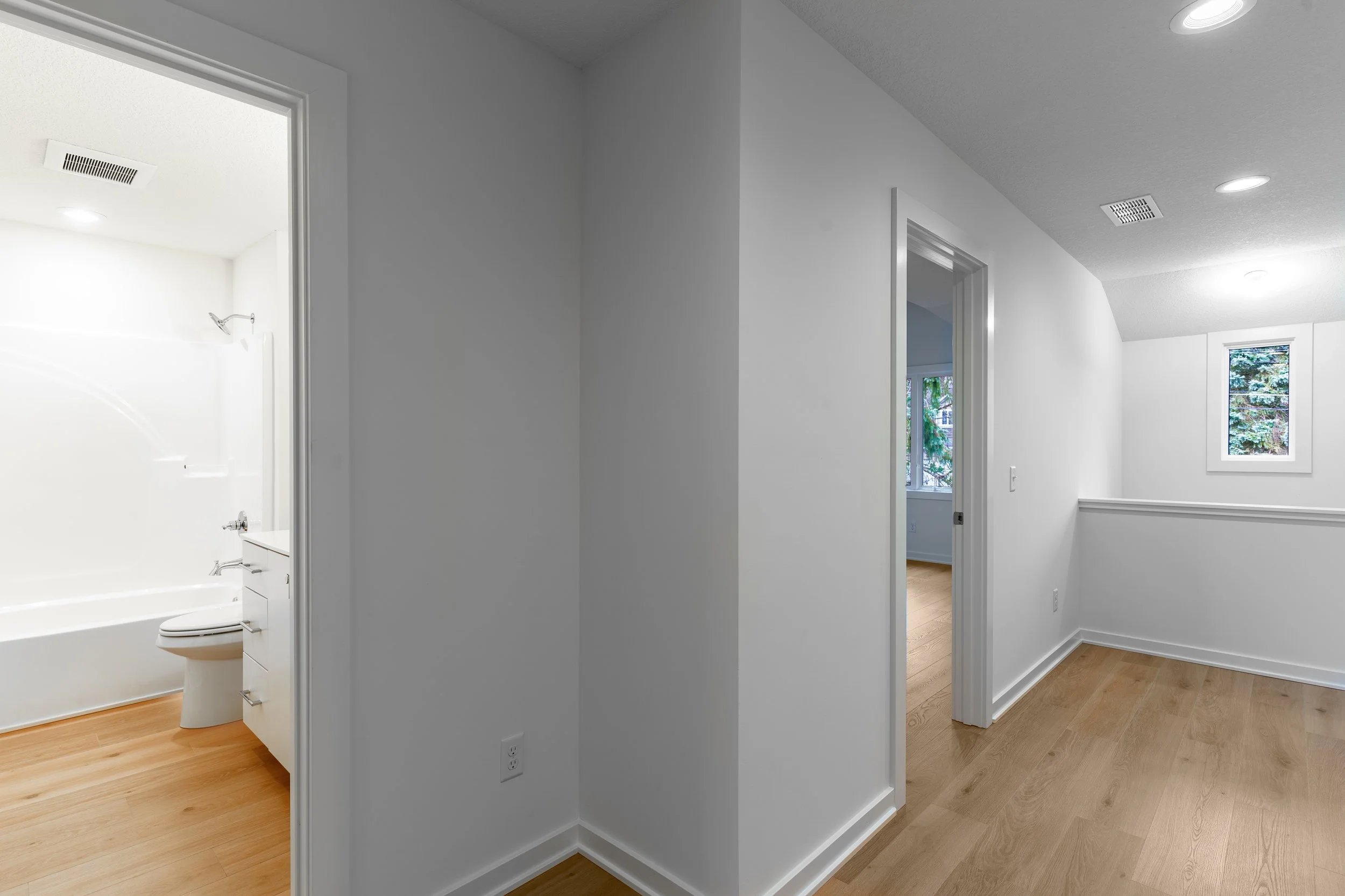 Interior view of a white-walled room with hardwood floors, an open doorway showing a bathroom, and a window with trees outside.