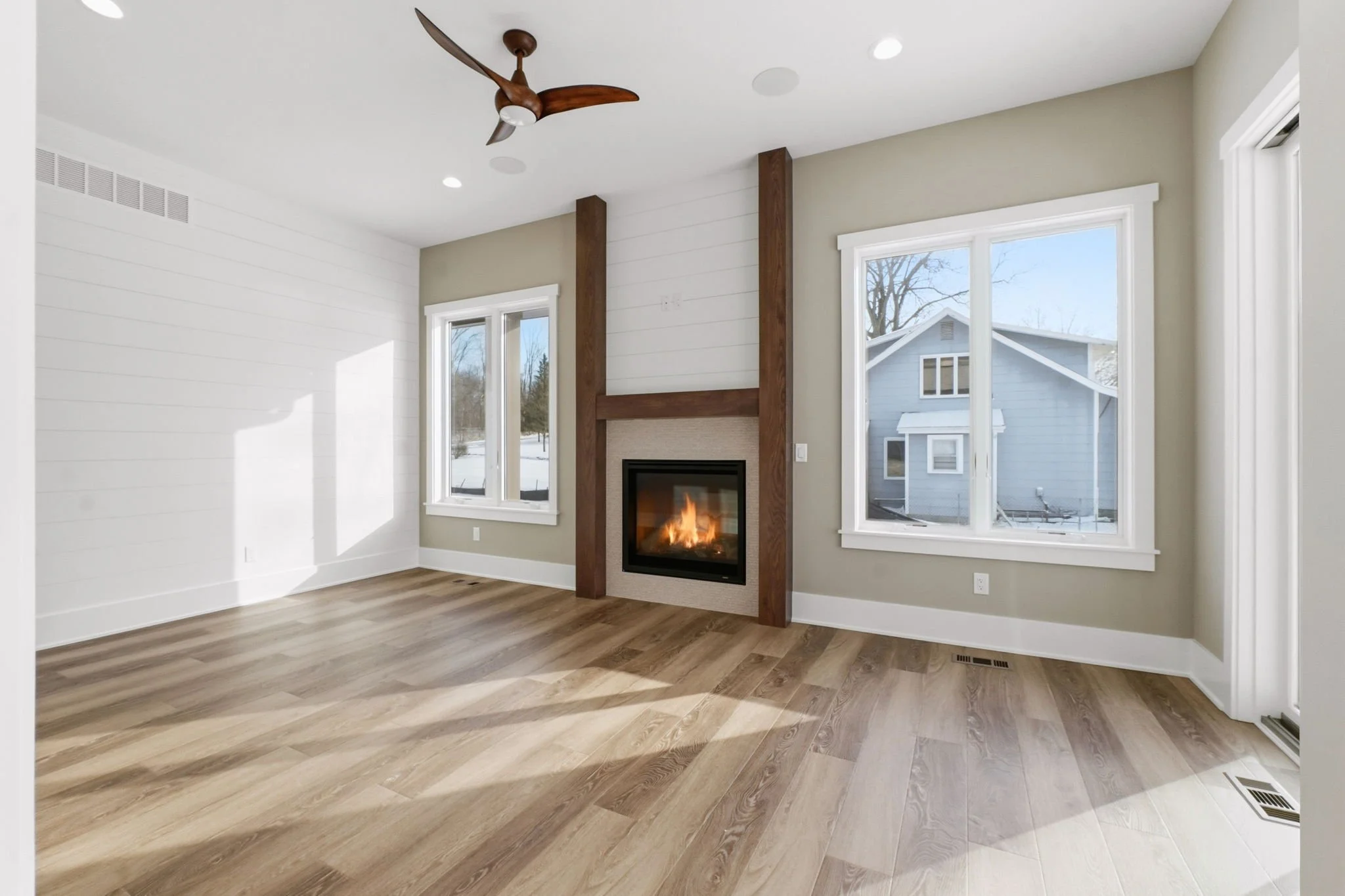 Empty living room with hardwood floors, a modern fireplace with wooden beams, large windows, and a ceiling fan.