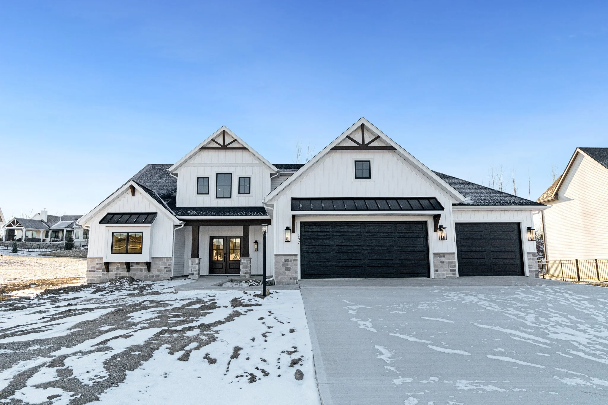 Modern white and black house with two garages, black doors, stone accents, black-lit exterior lamps, snow-covered front yard and driveway, and neighboring houses visible in the background under a blue sky.