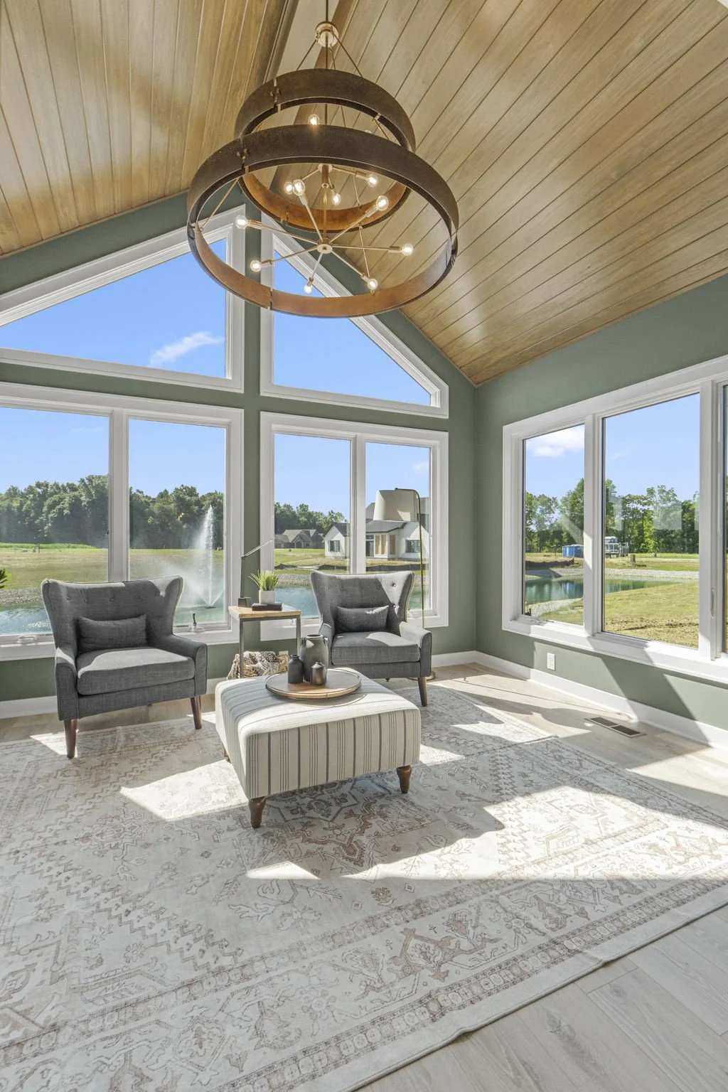 Sunlit living room with large windows overlooking a pond and trees, two gray armchairs with cushions, a striped ottoman with decorative items, a side table with a plant, and a wooden ceiling with a modern chandelier.