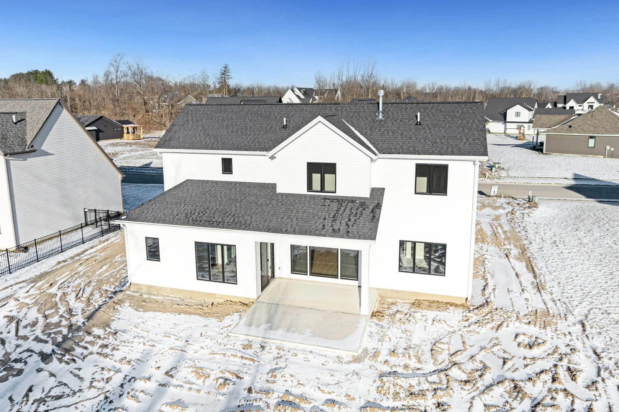 A modern two-story house with white exterior walls and a gray shingled roof, surrounded by a snow-covered yard and neighboring houses in a suburban neighborhood under a clear blue sky.