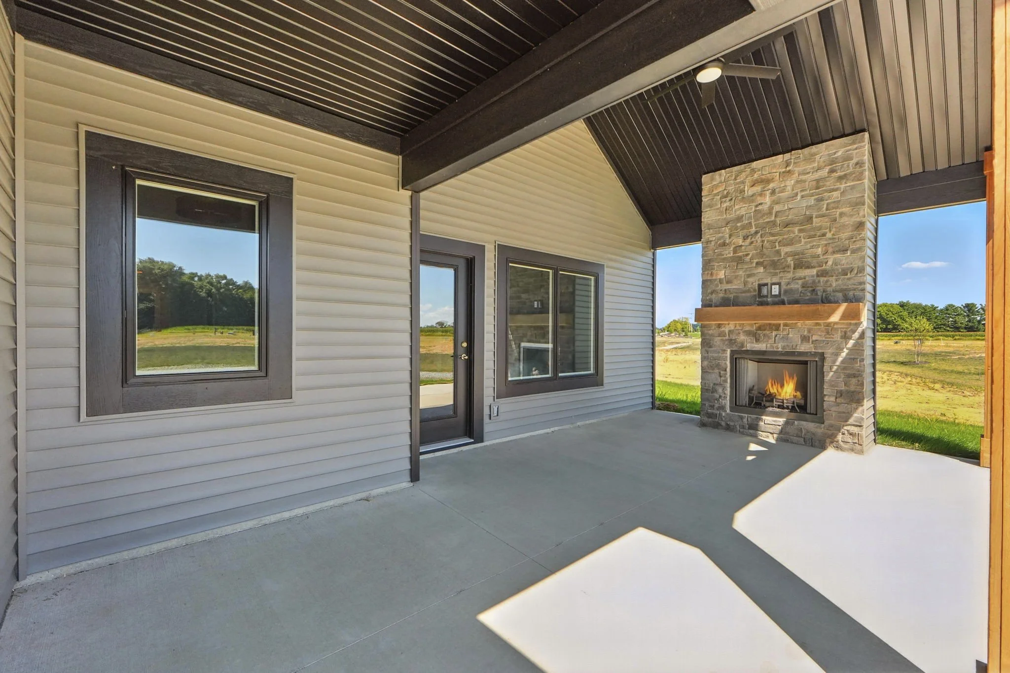 Covered patio area with concrete floor, beige siding walls with dark brown trim, a stone fireplace with a wooden mantel, and a view of a grassy field and trees in the background.