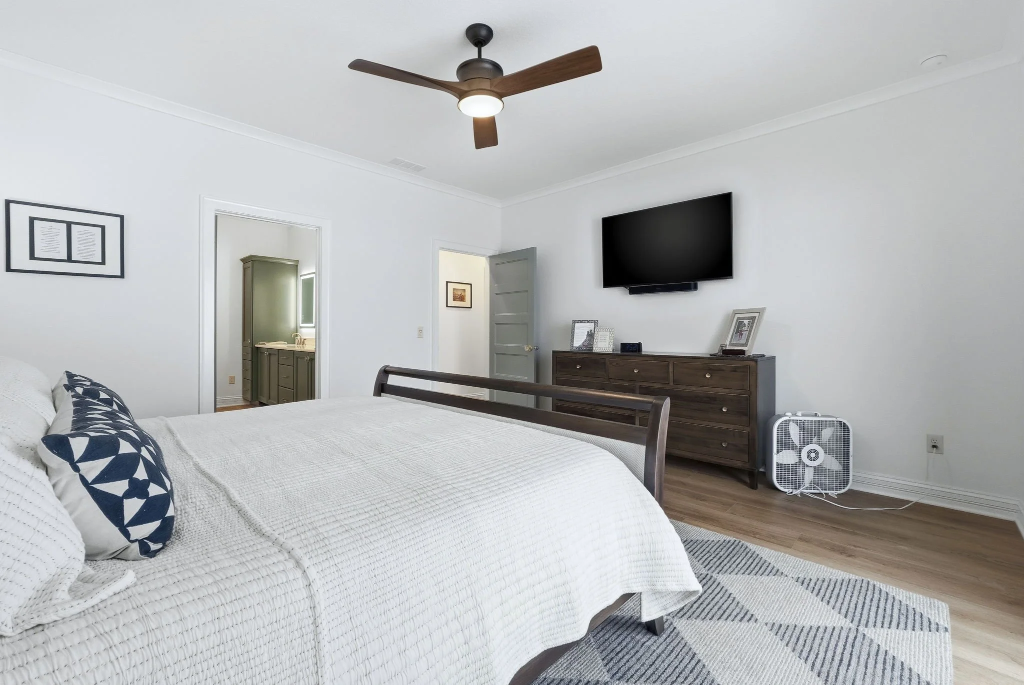 A bedroom with a large bed, white bedding, navy blue and white pillows, a wooden dresser with picture frames, a wall-mounted TV, a ceiling fan with wooden blades, a small fan on the floor, and a doorway leading to a bathroom.