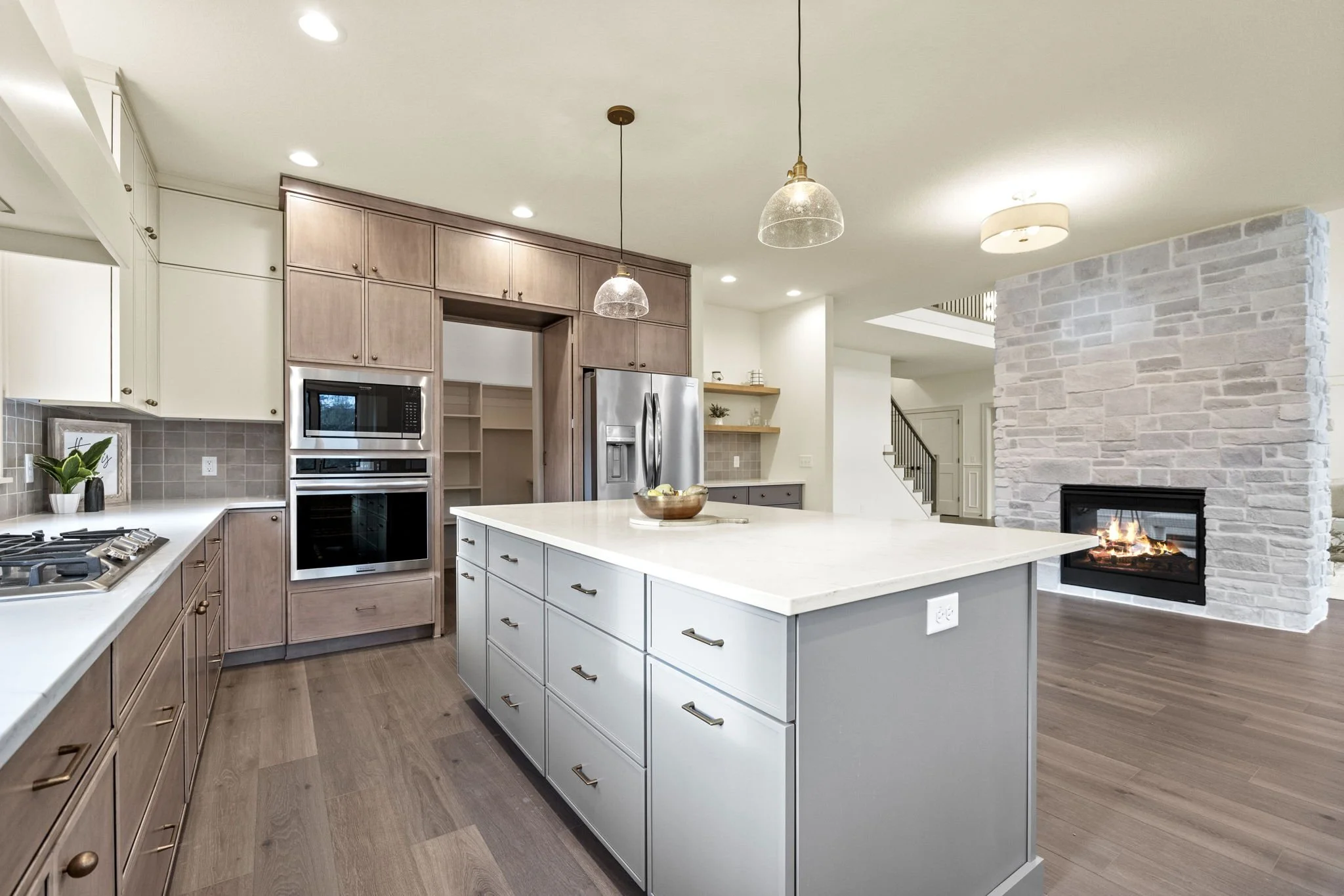 Modern kitchen with white and gray cabinetry, a large kitchen island with drawers, stainless steel appliances including a refrigerator and built-in oven, a gas stove, pendant lights, and a stone fireplace with a fire burning in an open living space.