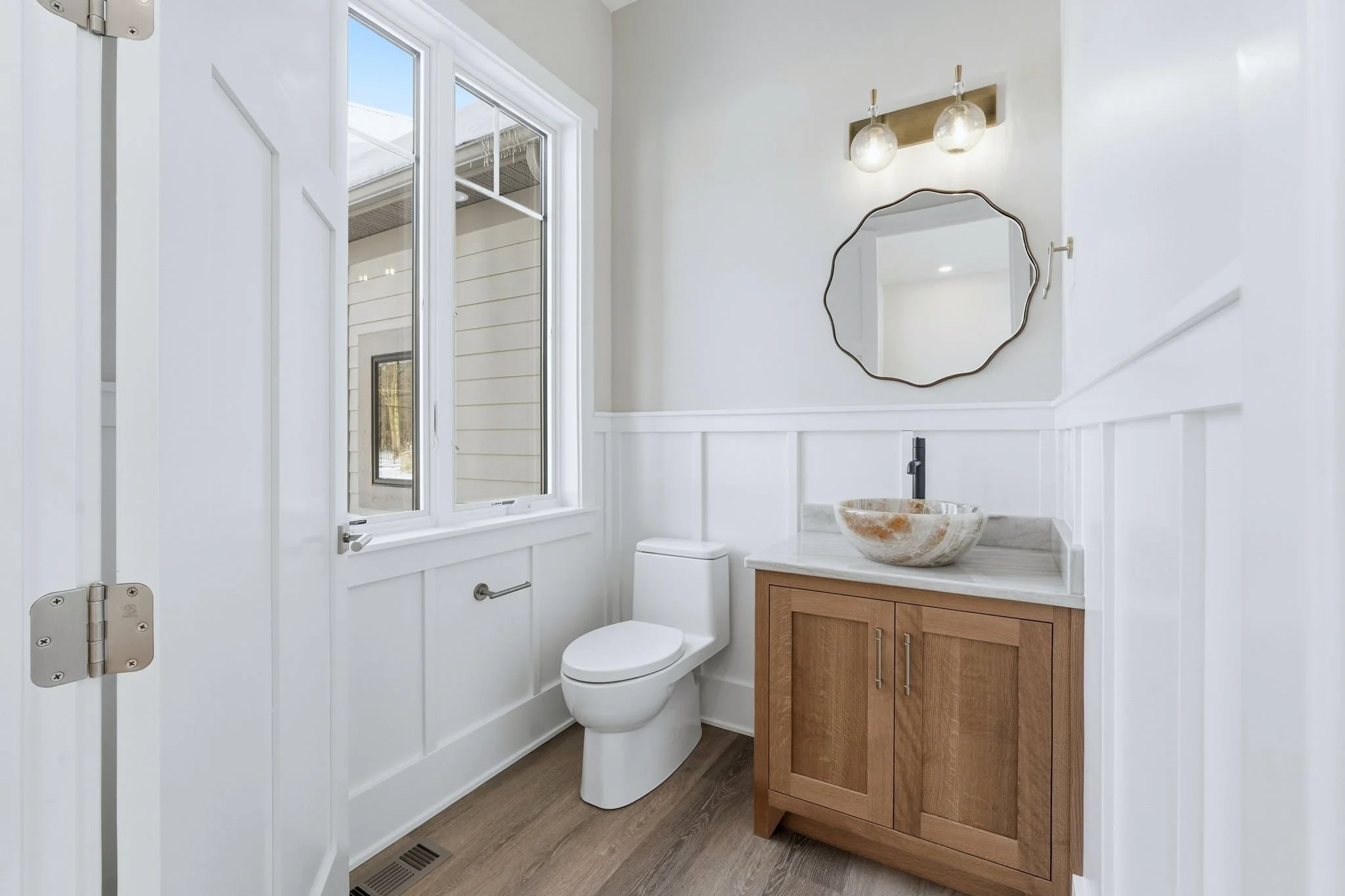 Modern bathroom with white walls, a toilet, a wooden vanity with a marble countertop, a vessel sink, and a small round mirror. There is a window letting in natural light and two wall-mounted light fixtures above the mirror.