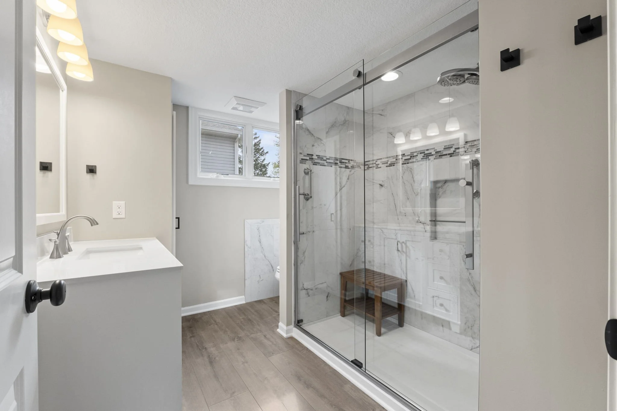 Modern bathroom with walk-in glass shower, marble tile walls, wooden stool, small window, and white vanity with sink and mirror.