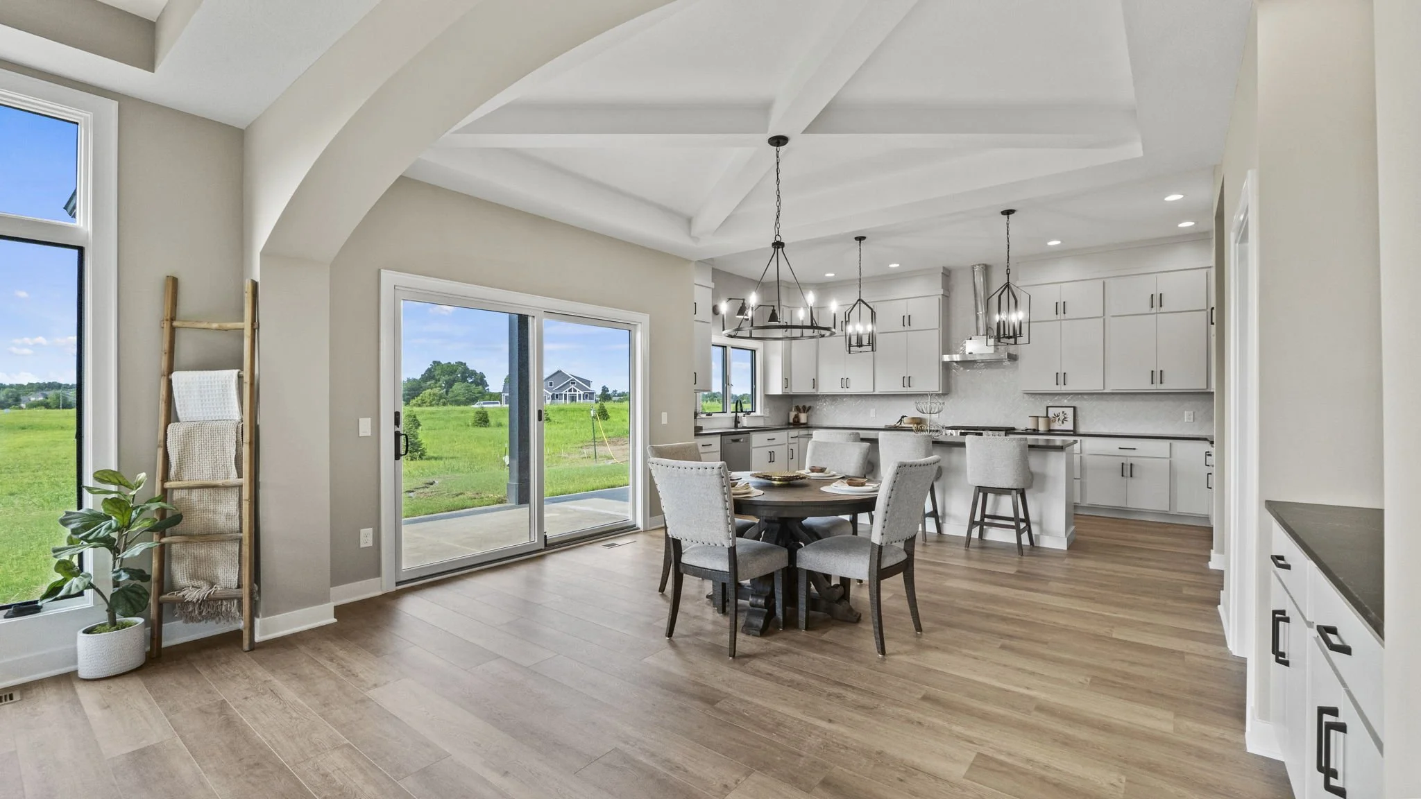 Open-concept kitchen and dining area with hardwood floors, white cabinetry, and large windows showing a green outdoor landscape.