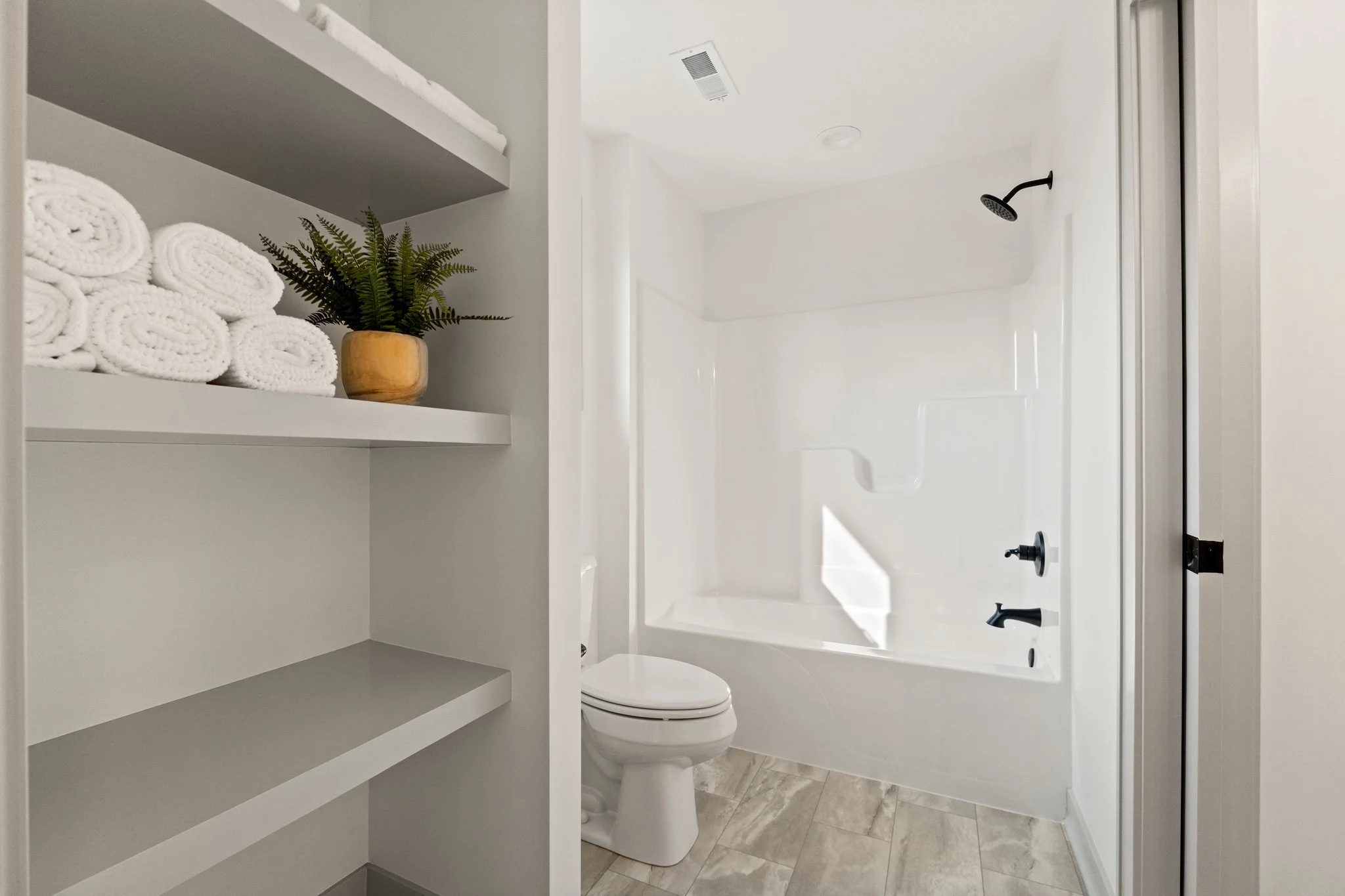 A bright bathroom with a white bathtub, black fixtures, a toilet, and shelves holding white towels and a potted plant.