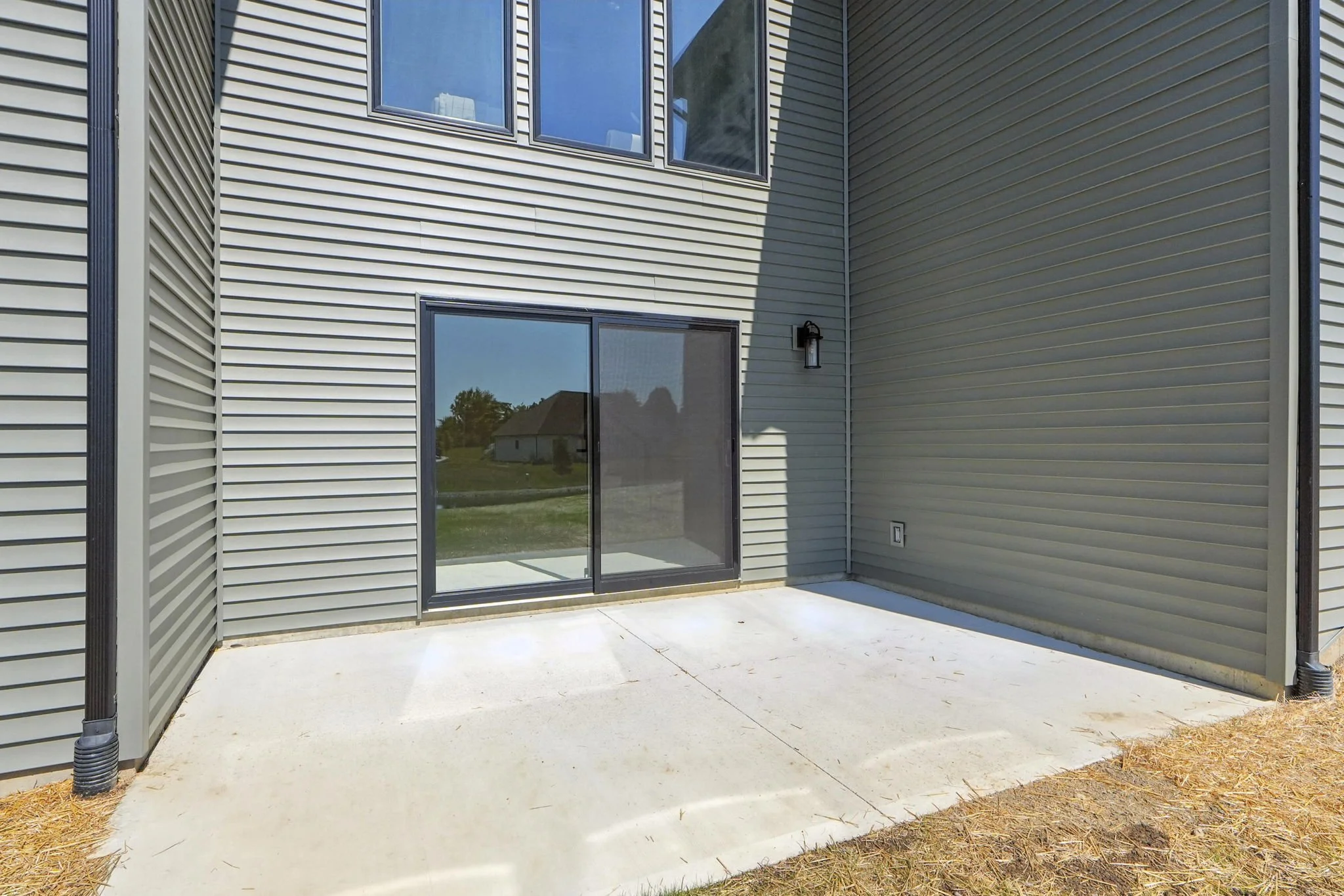 Empty backyard patio with concrete floor and sliding glass door leading into a modern house with beige siding and large windows.