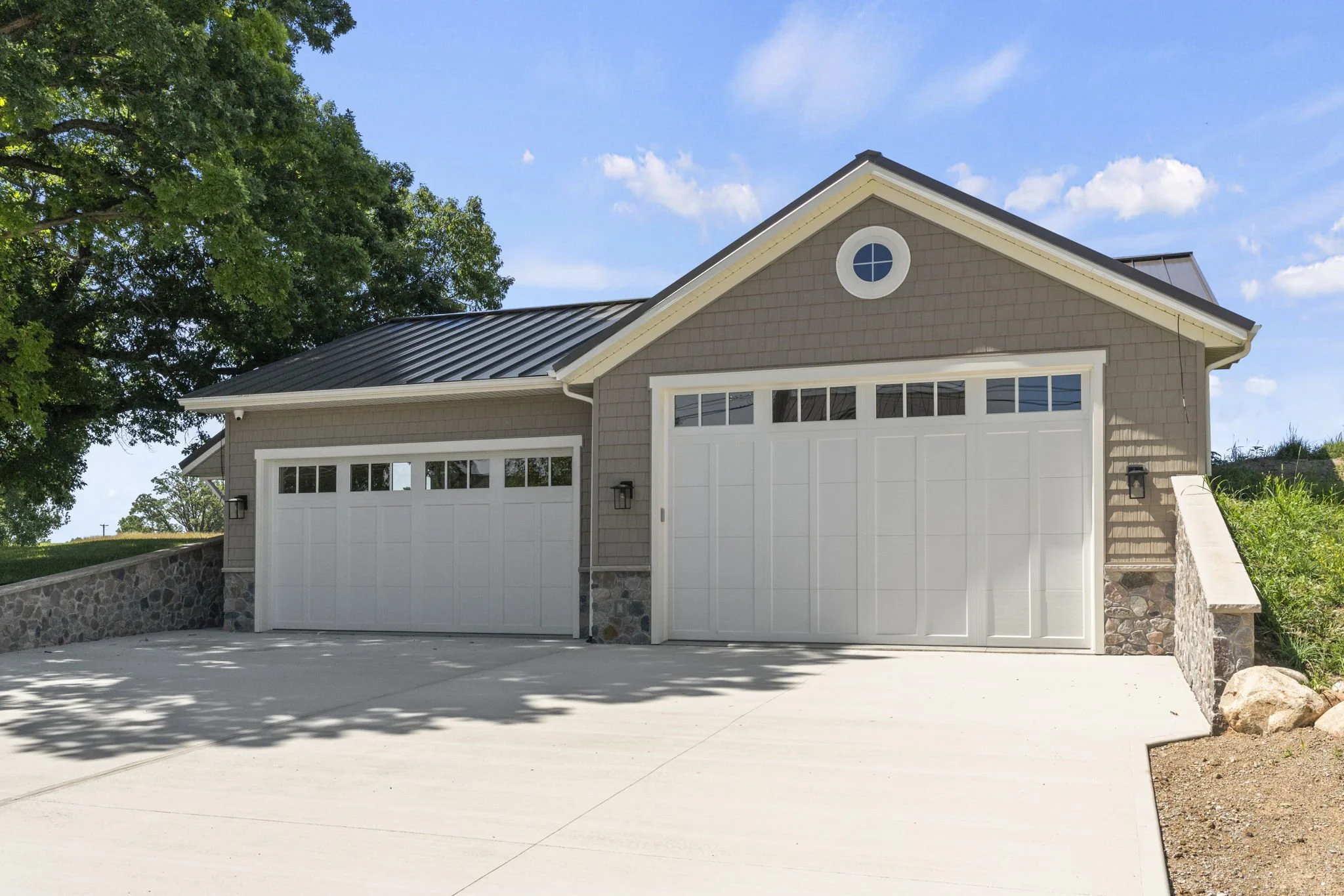 Front of a residential house with a double garage, stone accents on the lower walls, a gable roof, and a small circular window, surrounded by a driveway, a tree on the left, and a blue sky with a few clouds.
