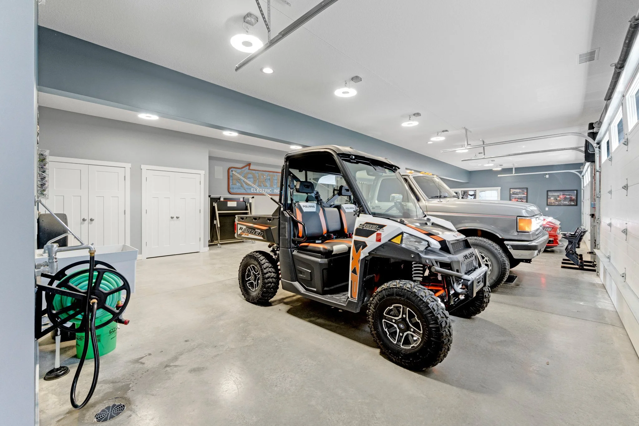 A spacious garage with three vehicles: a small off-road utility vehicle in the foreground, a pickup truck in the middle, and part of a red sports car in the background. The garage has a concrete floor, white walls, and overhead lighting. On the left,