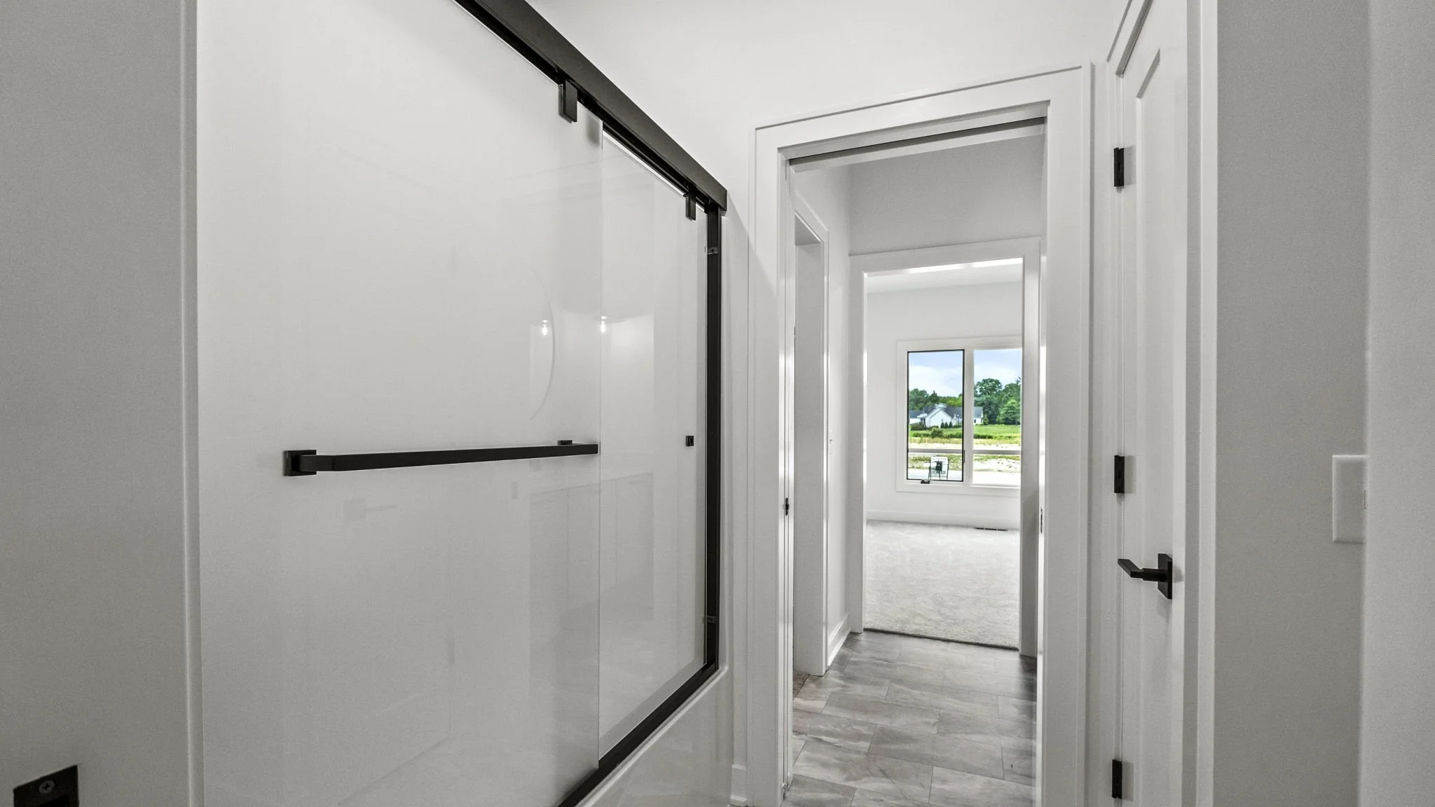 Inside view of a modern, bright room with a large window showing greenery outside, seen through a doorway with white walls and gray flooring.