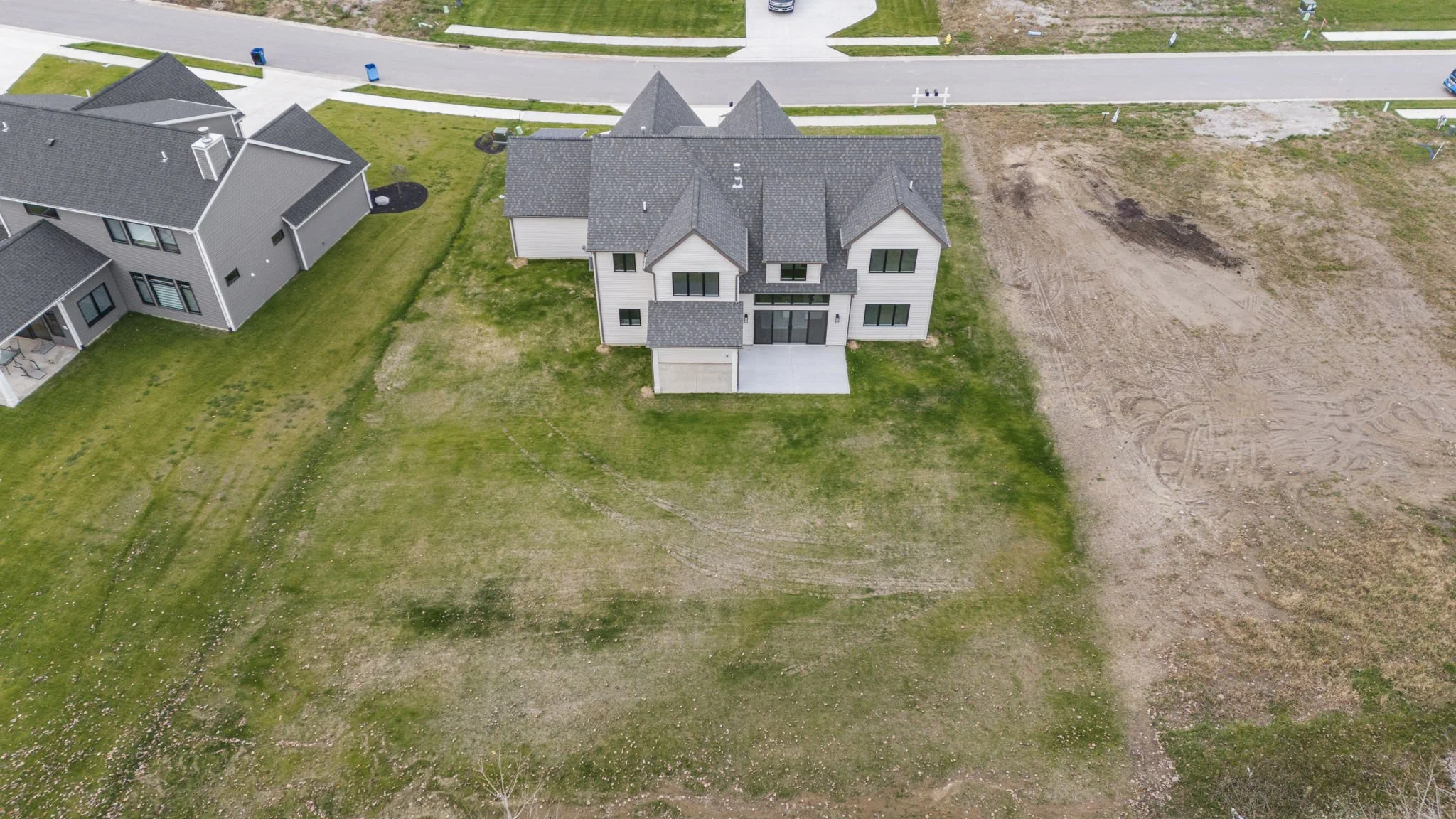 Aerial view of a newly constructed two-story house with a gray roof and white siding, adjacent to other similar houses, with a sidewalk and empty lot around it.