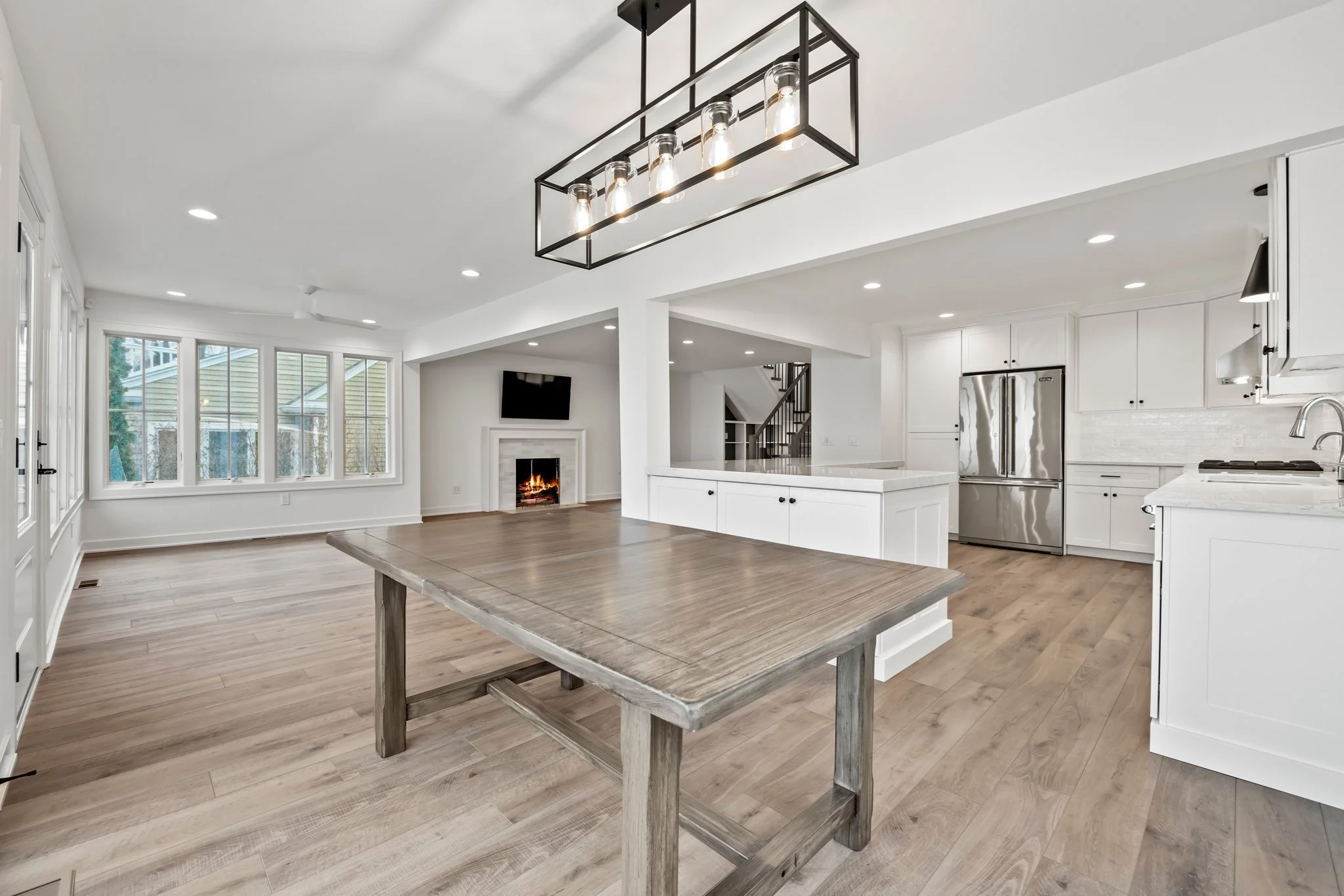 Empty open-concept kitchen and living room with wooden flooring, white cabinetry, stainless steel refrigerator, large kitchen island, and a fireplace with a wall-mounted TV above, illuminated by a modern rectangular light fixture.