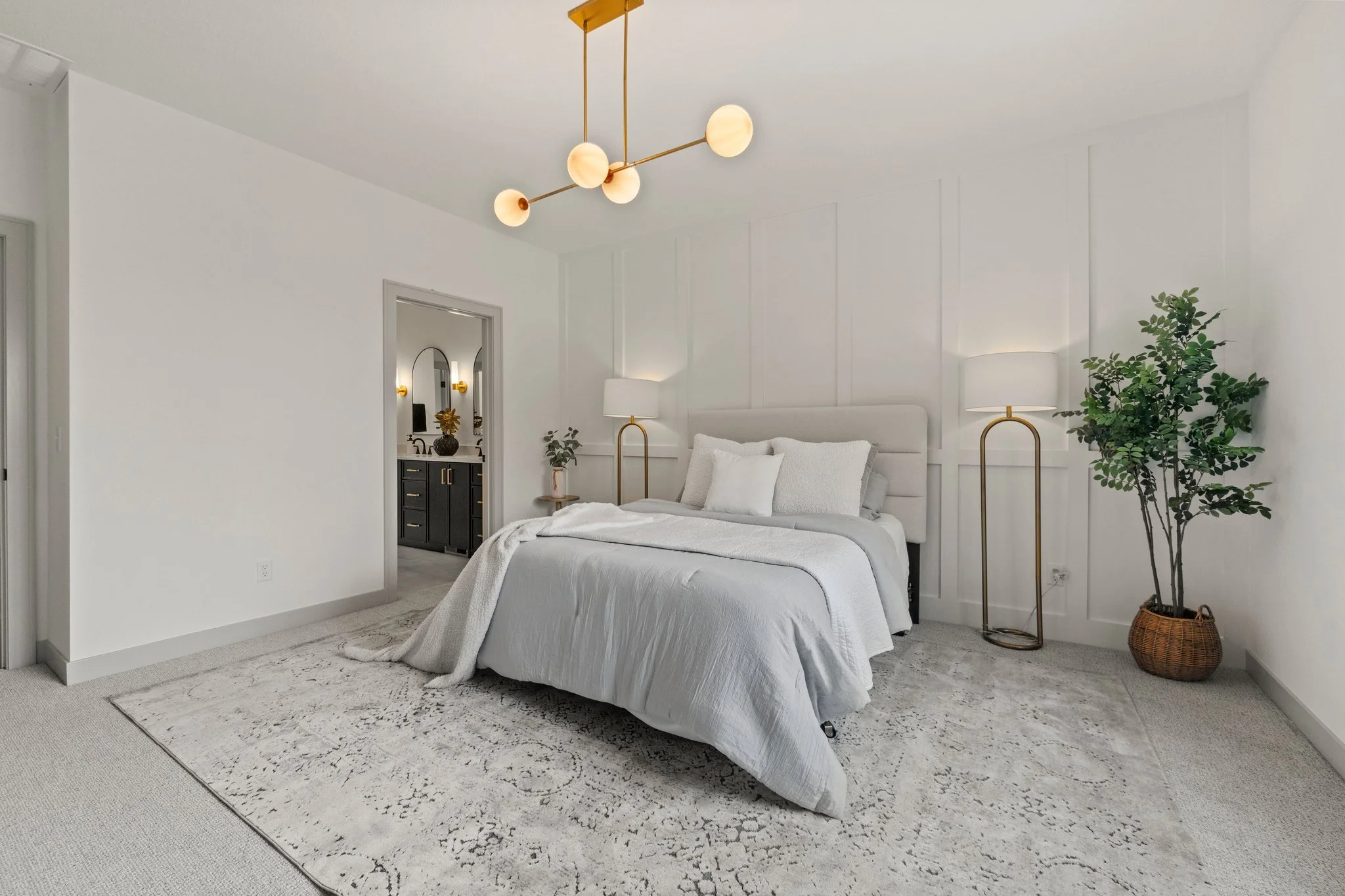 A modern bedroom with white walls and a white bed. Bedside lamps and a large potted plant are present, with a black dresser with gold handles and a mirror reflecting light fixtures visible through an open door.