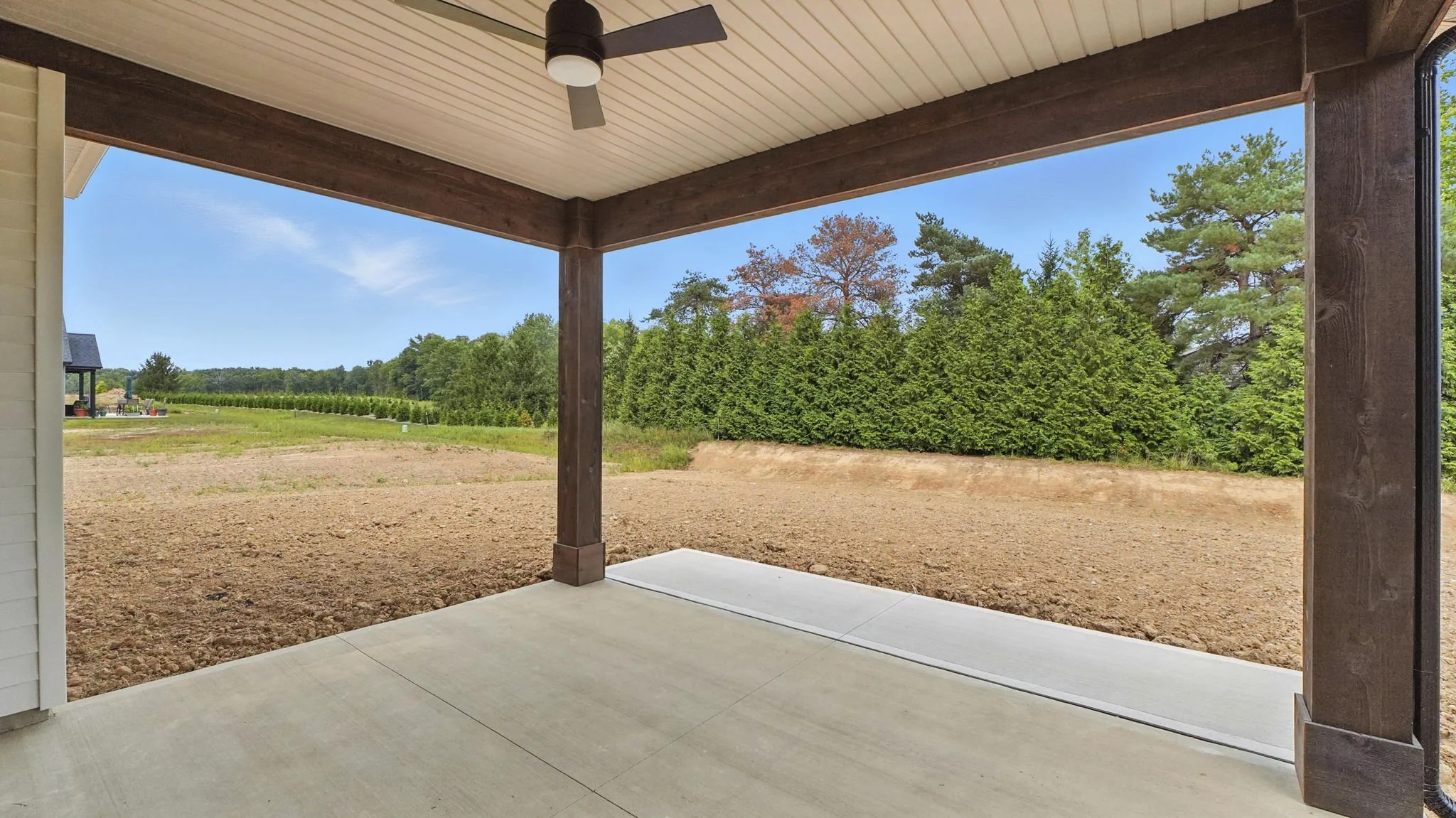 View from a covered patio looking out to a backyard with dirt ground and a row of tall green trees and a blue sky.