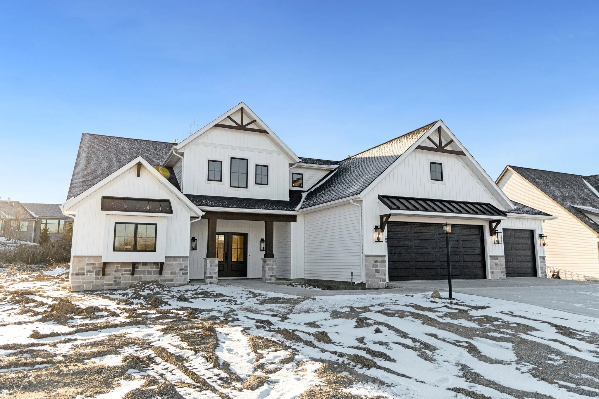 Modern white house with black accents, a two-car garage, and a front porch with columns, snow on the ground, and a clear blue sky.