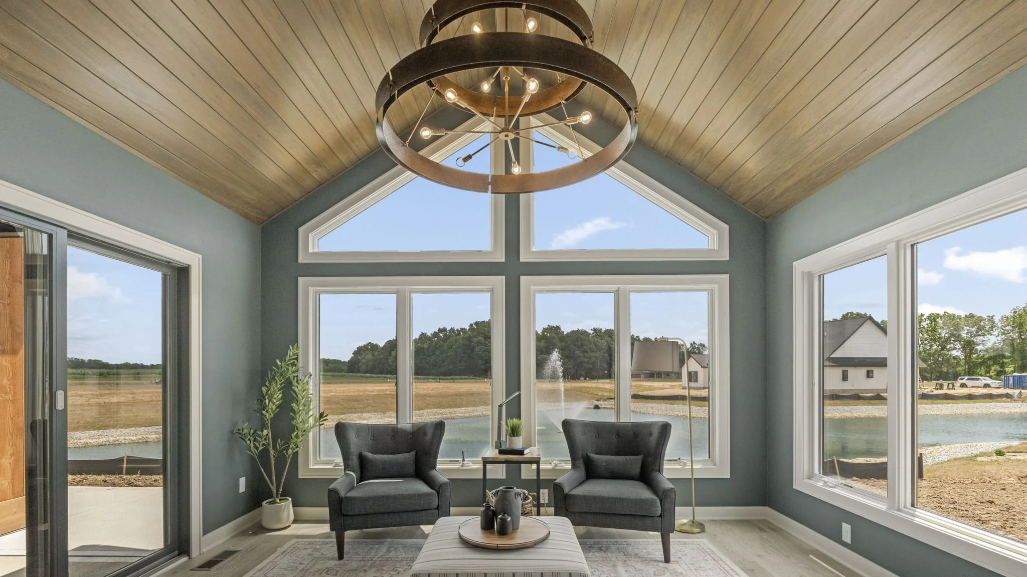 Living room with large windows overlooking a pond, with two dark gray armchairs and a small table between them, a house and trees outside, a modern chandelier hanging from a wooden ceiling.