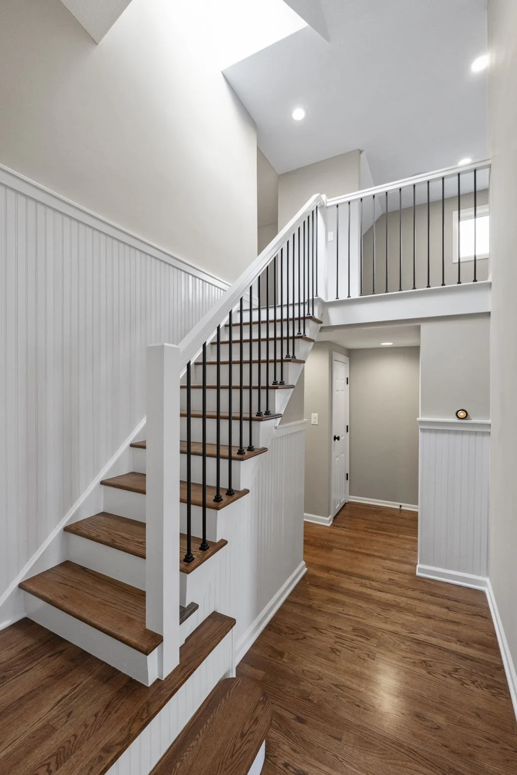 Interior view of a staircase with wooden steps, white risers, black iron balusters, and white walls with beadboard paneling, leading to an upper floor, with a small landing and a hallway at the bottom.