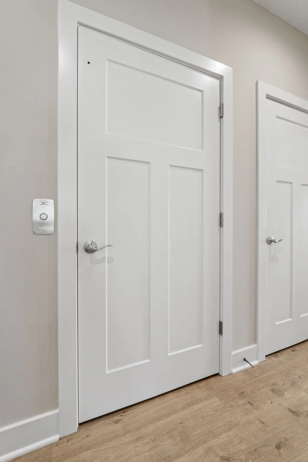 White interior door with silver handle and hinges, beige walls, light wood floor, and a thermostat on the wall to the left.