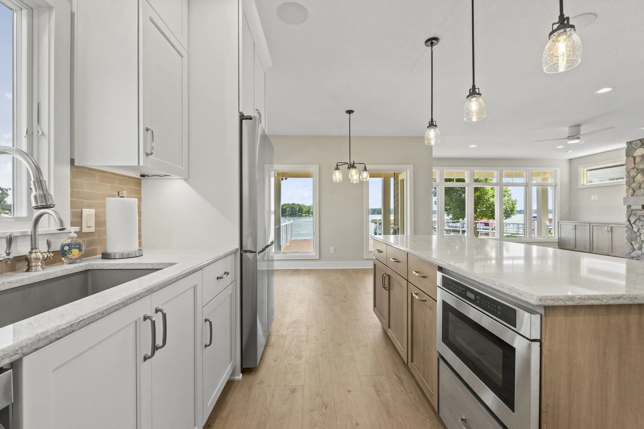 Modern kitchen with white cabinets, light wood flooring, stainless steel appliances, and large windows overlooking water with boats and trees.