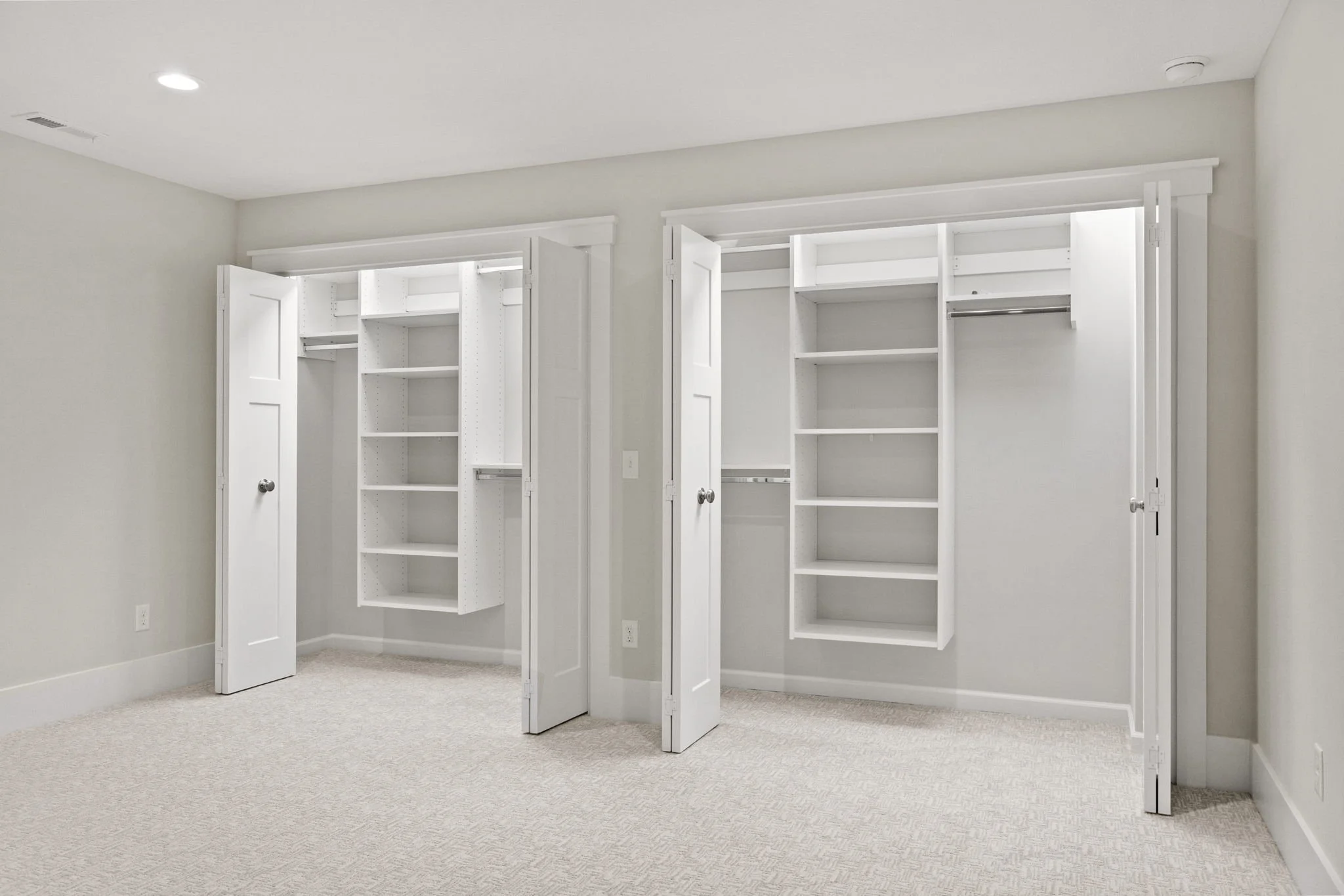 Empty walk-in closets with white shelves and hanging rods in a room with beige carpet and white walls.