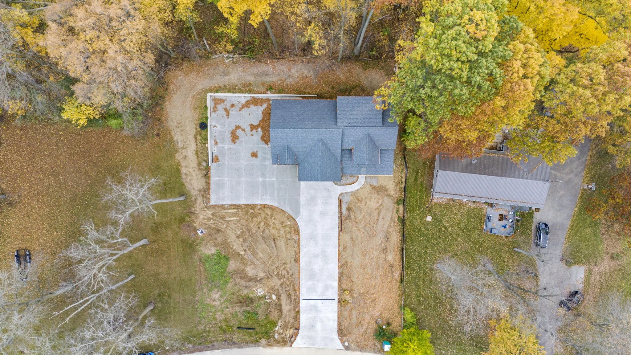 An aerial view of a house with a blue roof, surrounded by trees with autumn foliage, a concrete driveway, and a neighboring building with a metal roof.