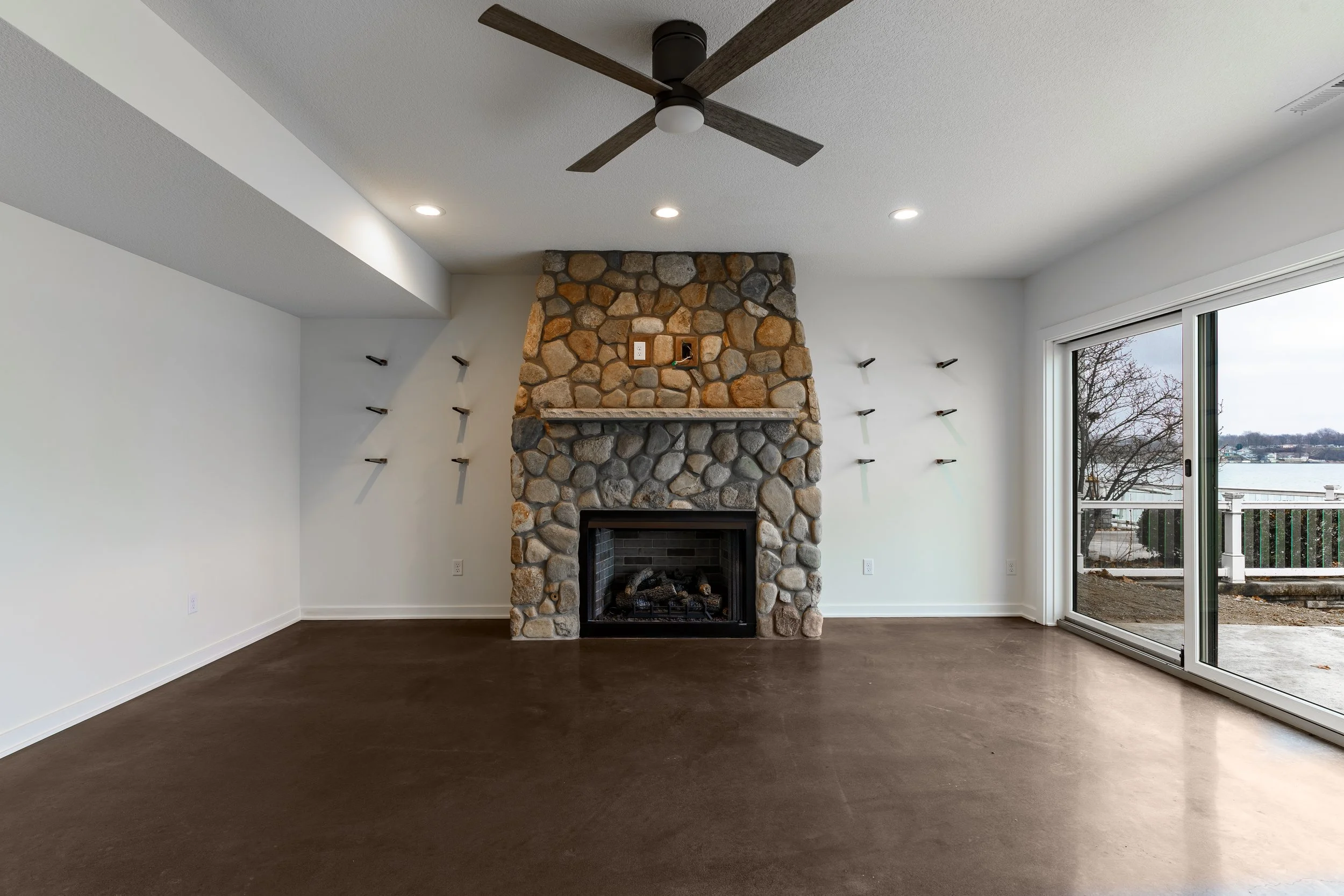 Empty living room with a stone fireplace, a ceiling fan, and sliding glass doors leading outside.
