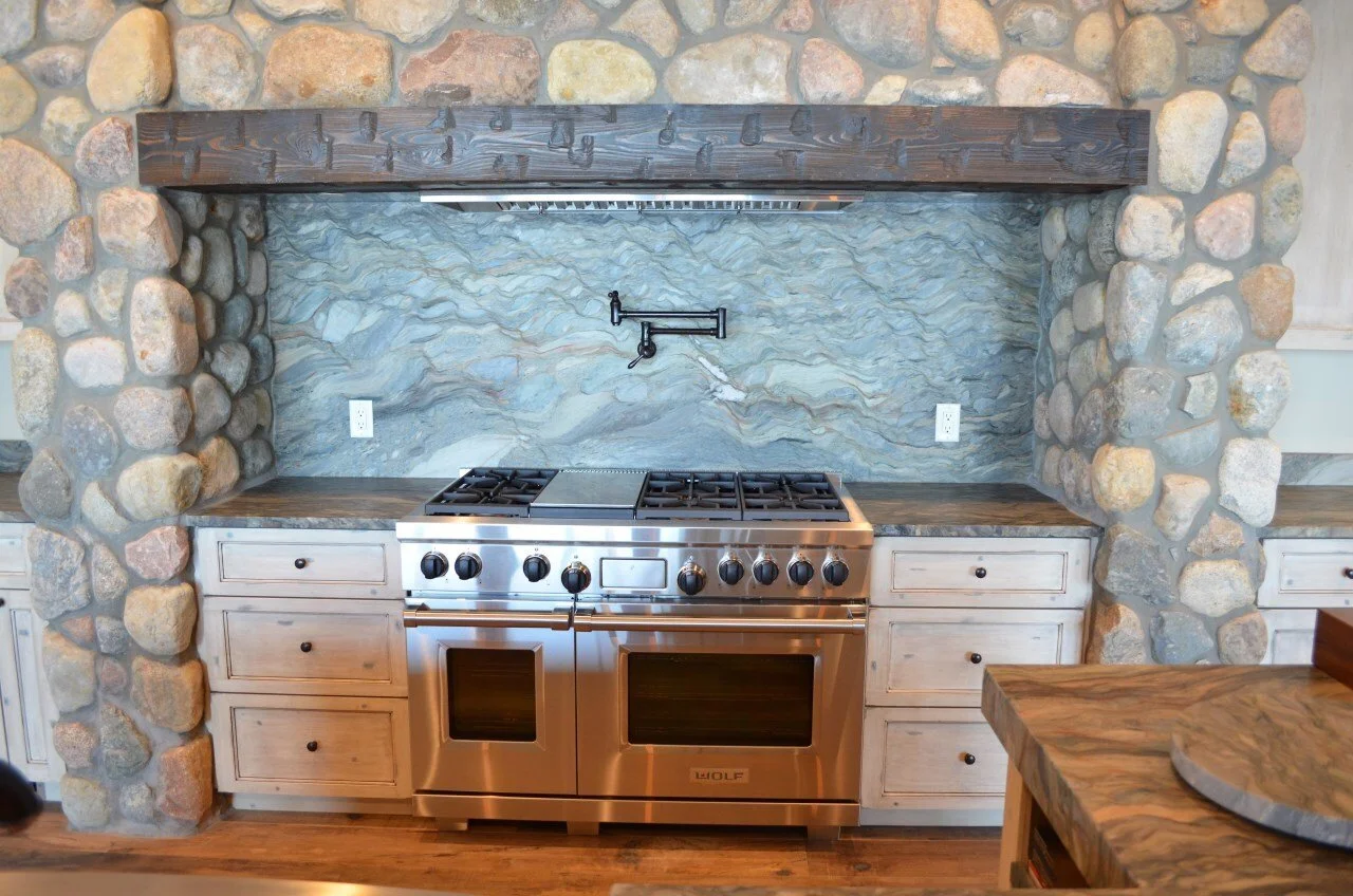 Kitchen with stone arch framing a stainless steel stove, dark wood countertops, white cabinetry, and a textured stone wall backdrop.