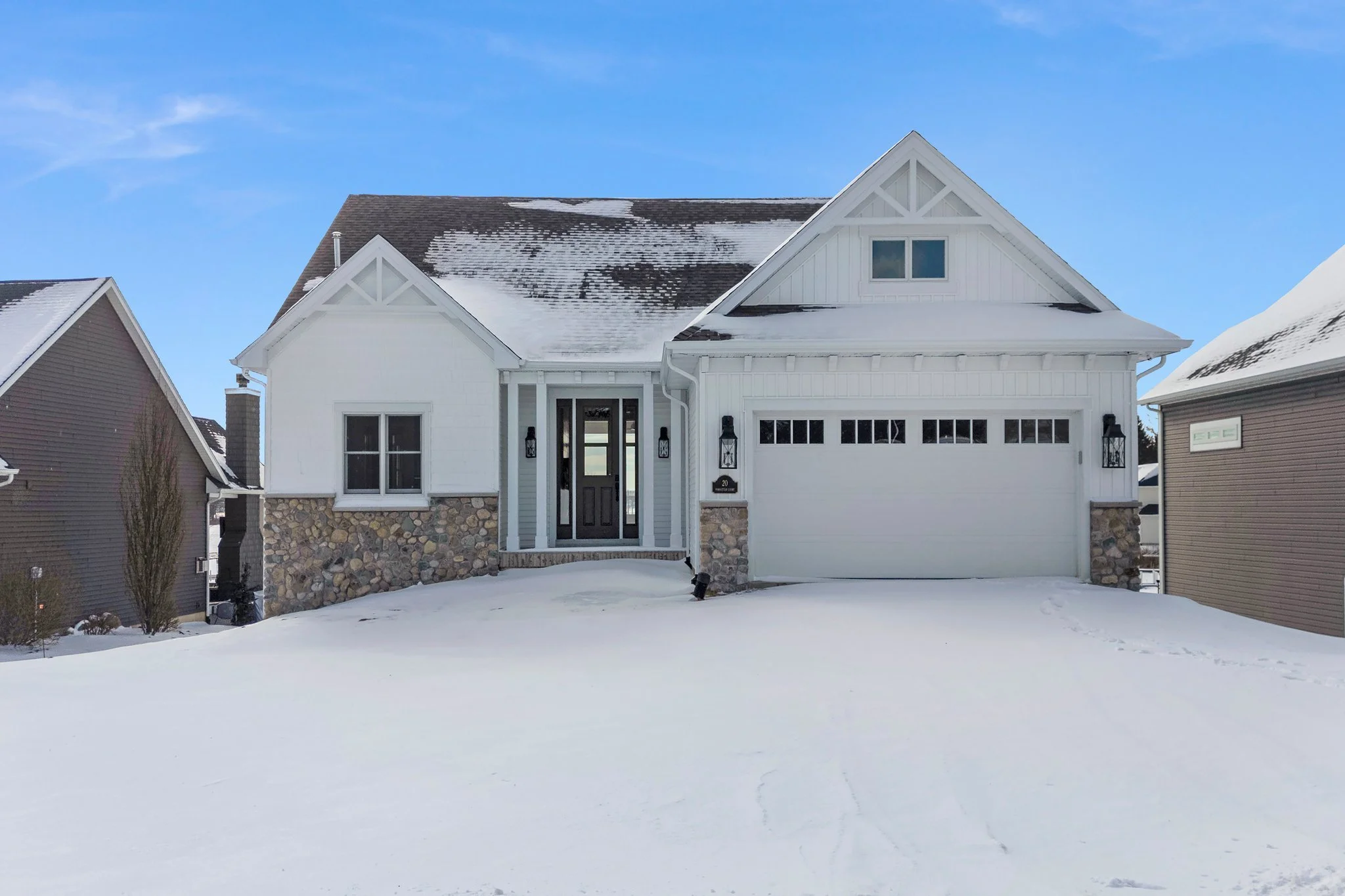 A white house with stone accents on the lower half, snow on the roof and ground, and blue sky in the background.
