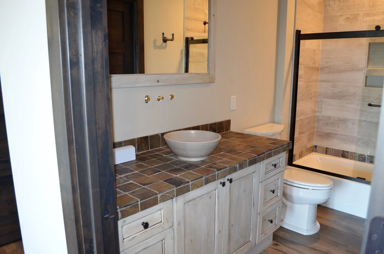 Bathroom with a vanity featuring a rustic tiled countertop, a vessel sink, and a large mirror. There is a toilet and a shower with a glass door, tile walls, and a small built-in shelf.