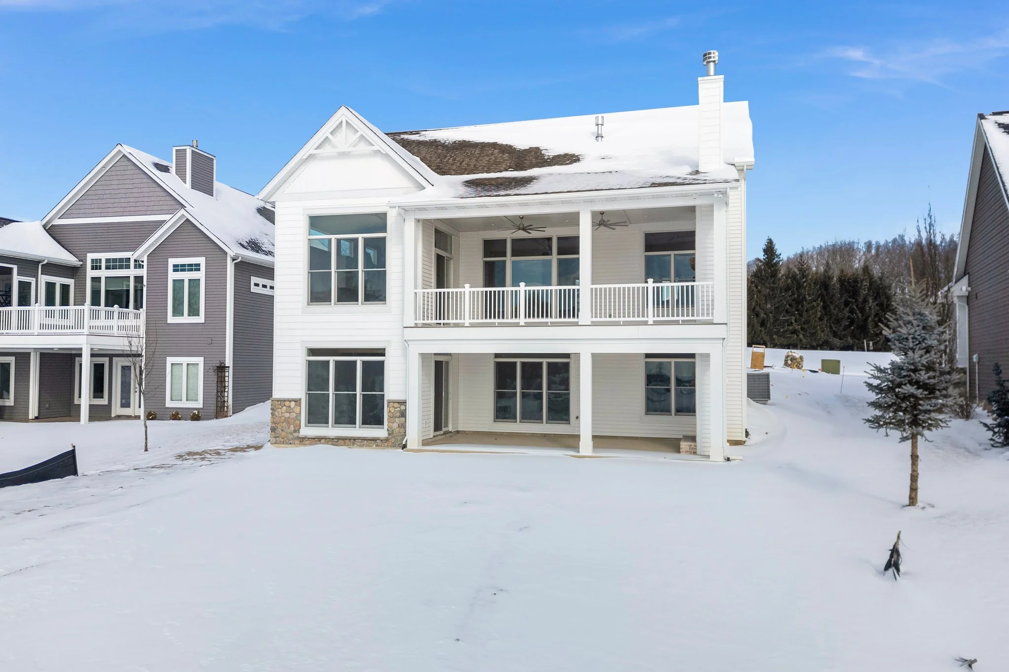 White two-story house with a covered back porch and upper balcony, snow-covered lawn and roof, neighboring houses, clear blue sky, small trees in the snow.